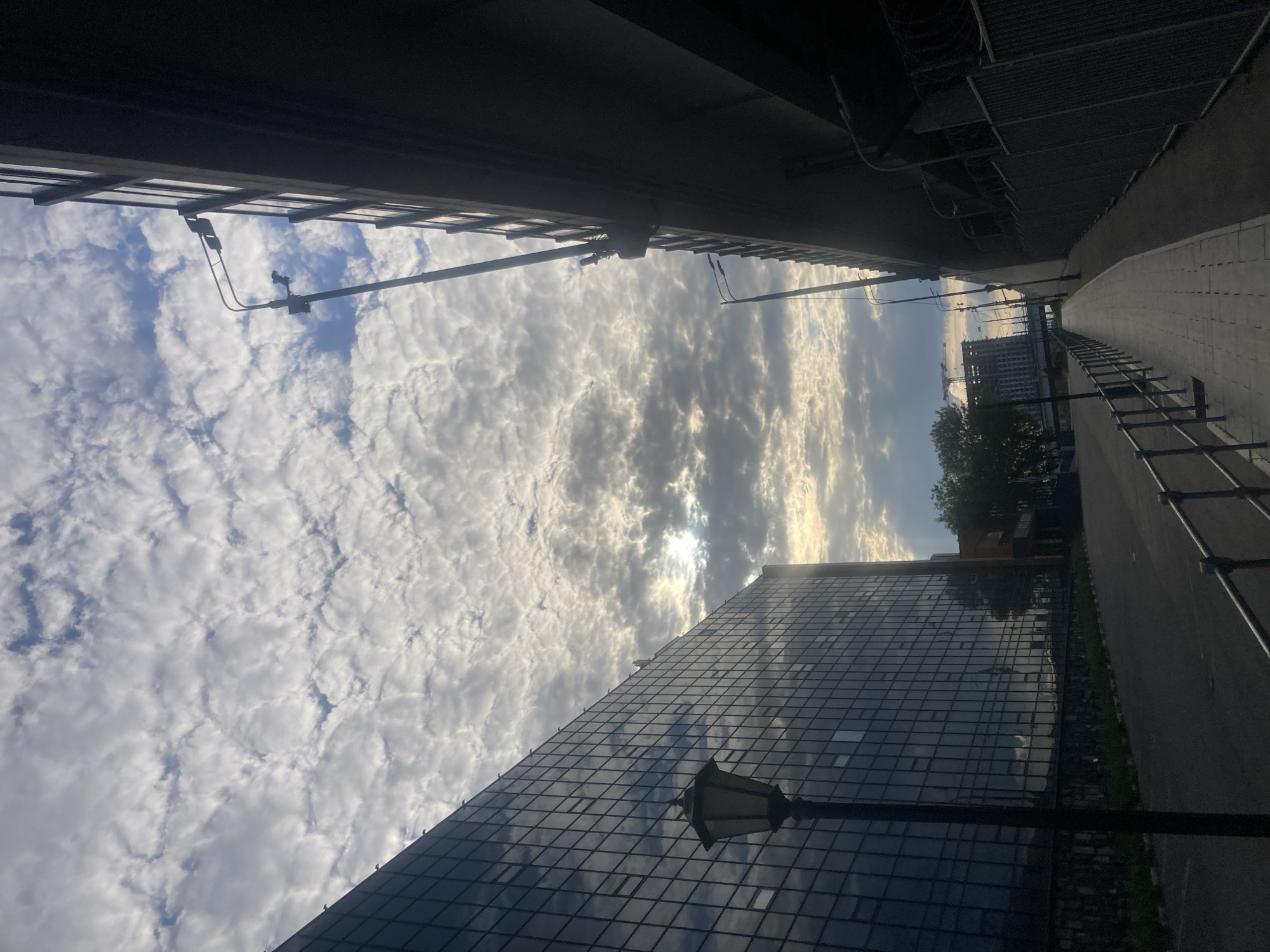 The image, rotated for easier viewing, depicts a street-level view in what appears to be a city, Moscow, Russia. The sky is filled with clouds, illuminated by sunlight. A modern, high-rise building with a reflective glass facade is visible on the right side of the frame. A street lamp is positioned near the bottom right corner. On the right, a sidewalk runs along the edge of the street, bordered by a metal railing. Overhead, there is an element of infrastructure such as an overpass or bridge. In the distance, buildings and construction cranes are visible.