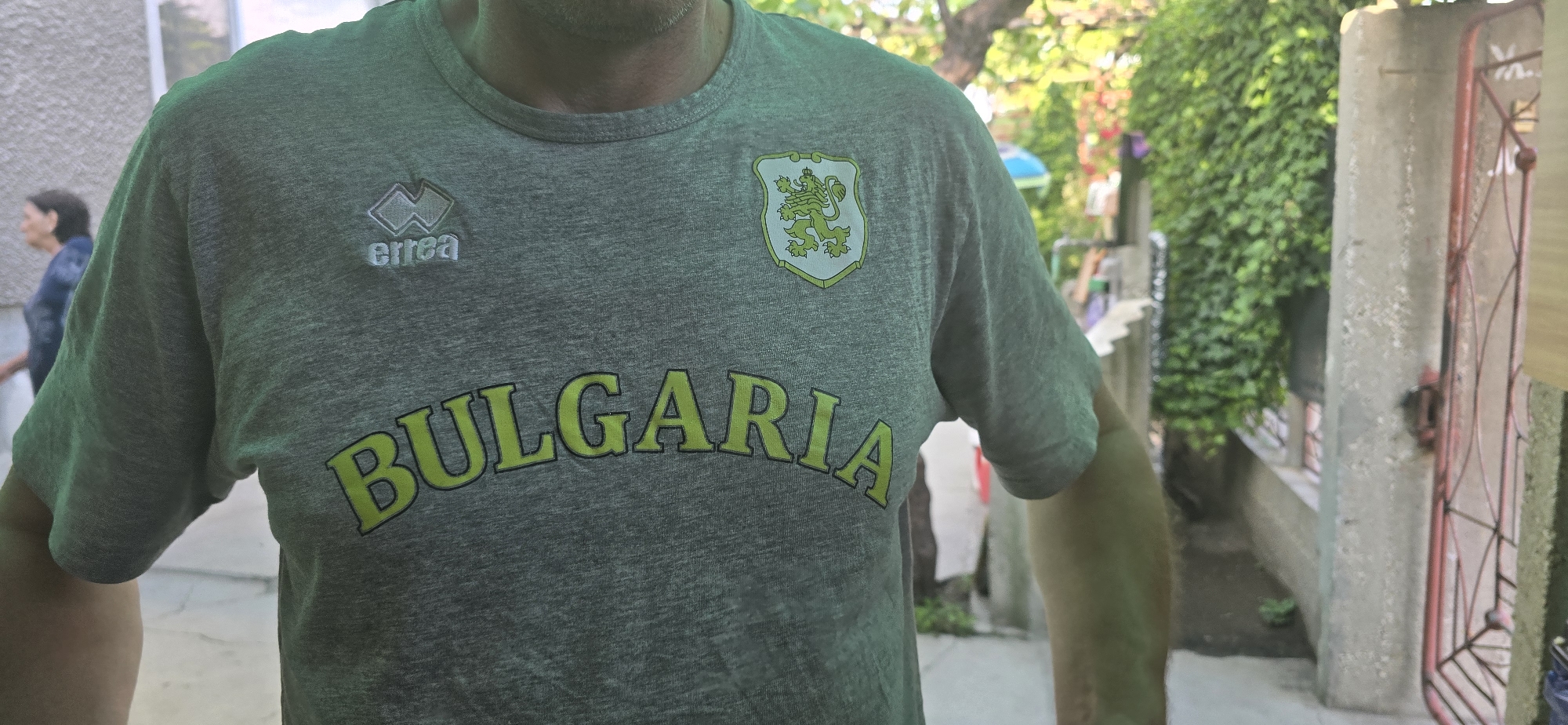 An adult individual, partially visible, stands outdoors in bright daylight. The main subject is wearing a heather grey t-shirt. On the upper left chest, the "Errea" logo is visible in white and grey. On the upper right chest, there is an emblem featuring a green crowned lion rampant on a shield, which is a common depiction of the coat of arms of Bulgaria. Below these, the word "BULGARIA" is prominently displayed across the chest in large, arched, yellow-green letters with a dark outline.
The background suggests a residential or garden setting. To the left, another person, a woman, is partially visible walking away. The environment features lush green foliage, likely ivy growing on a wall or fence, and some trees. On the right side of the frame, there's a concrete pillar and a red metal gate with intricate patterns. The ground appears to be a paved or concrete surface. The strong lighting indicates it is daytime, likely morning or afternoon, with dappled sunlight. The overall scene conveys a casual outdoor moment in what is identified as Govedare, Bulgaria.