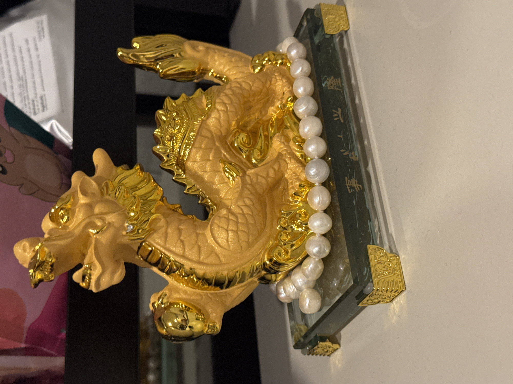 A gold-colored figurine, resembling a mythical creature such as a Pixiu or dragon, is prominently displayed in an indoor setting, likely in Bangkok, Thailand. The creature features intricate scales, a detailed head with a mane, and flowing tail, all rendered in a bright, shiny gold finish with some areas showing a slightly matte, textured gold. A string of irregularly shaped, lustrous white pearls is draped elegantly over the back of the creature and extends onto its base.
The figurine rests on a transparent, rectangular base, possibly made of glass or acrylic. This base is adorned with small, ornate golden corner brackets. Visible on the side of the transparent base are vertical characters written in an East Asian script, which appear to be Chinese. The characters "招財" (Zhao Cai - attracting wealth) and "進寶" (Jin Bao - bringing in treasure) are discernible, forming the common auspicious phrase "招財進寶" (Zhāo Cái Jìn Bǎo), wishing for wealth and prosperity.
In the background, a dark, vertical surface, possibly a piece of furniture or a wall, is visible. To the far left, a soft pink or peach-colored object with a partial cartoon illustration peeks into the frame. The immediate background to the right of the base is a plain, light-colored wall or surface. The overall impression is one of a decorative and culturally significant item, likely used for good fortune. No activity is taking place, and the time of day is not discernible.