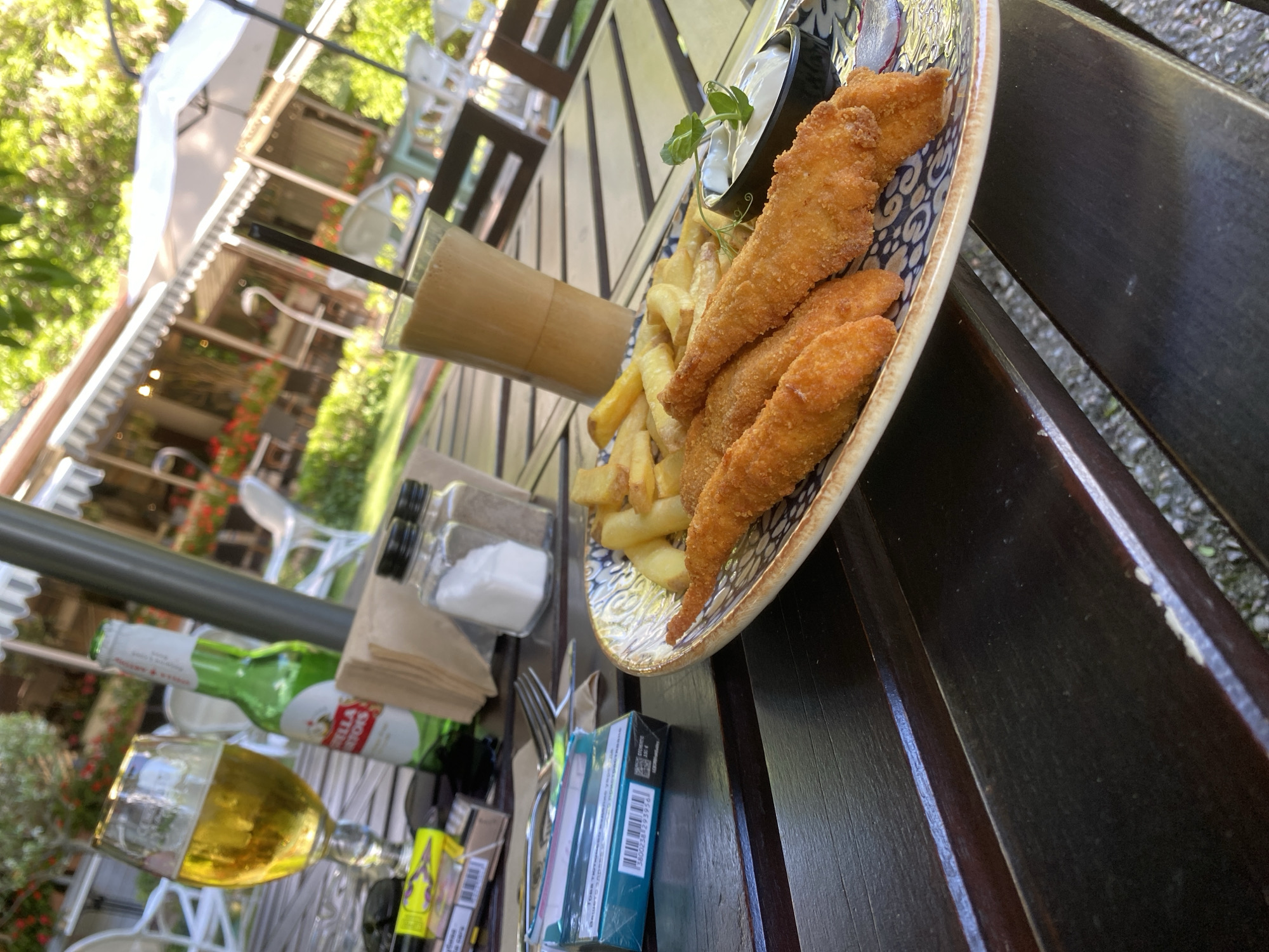 The image captures a close-up view of a wooden table at an outdoor restaurant, likely in Varna, Bulgaria, given the context. The table is set with a meal consisting of fried chicken strips and french fries, served on a patterned plate. To the left, a glass of beer and a green bottle of Stella Artois beer are visible, alongside a pack of cigarettes and a lighter. A tall glass of iced coffee with a straw stands in the background. The restaurant's outdoor seating area, with white chairs and tables partially covered by umbrellas and awnings, is discernible behind the table. Lush green foliage and trees suggest a pleasant, possibly warm-weather setting. The overall scene depicts a casual dining experience, with food and drinks laid out on the table.