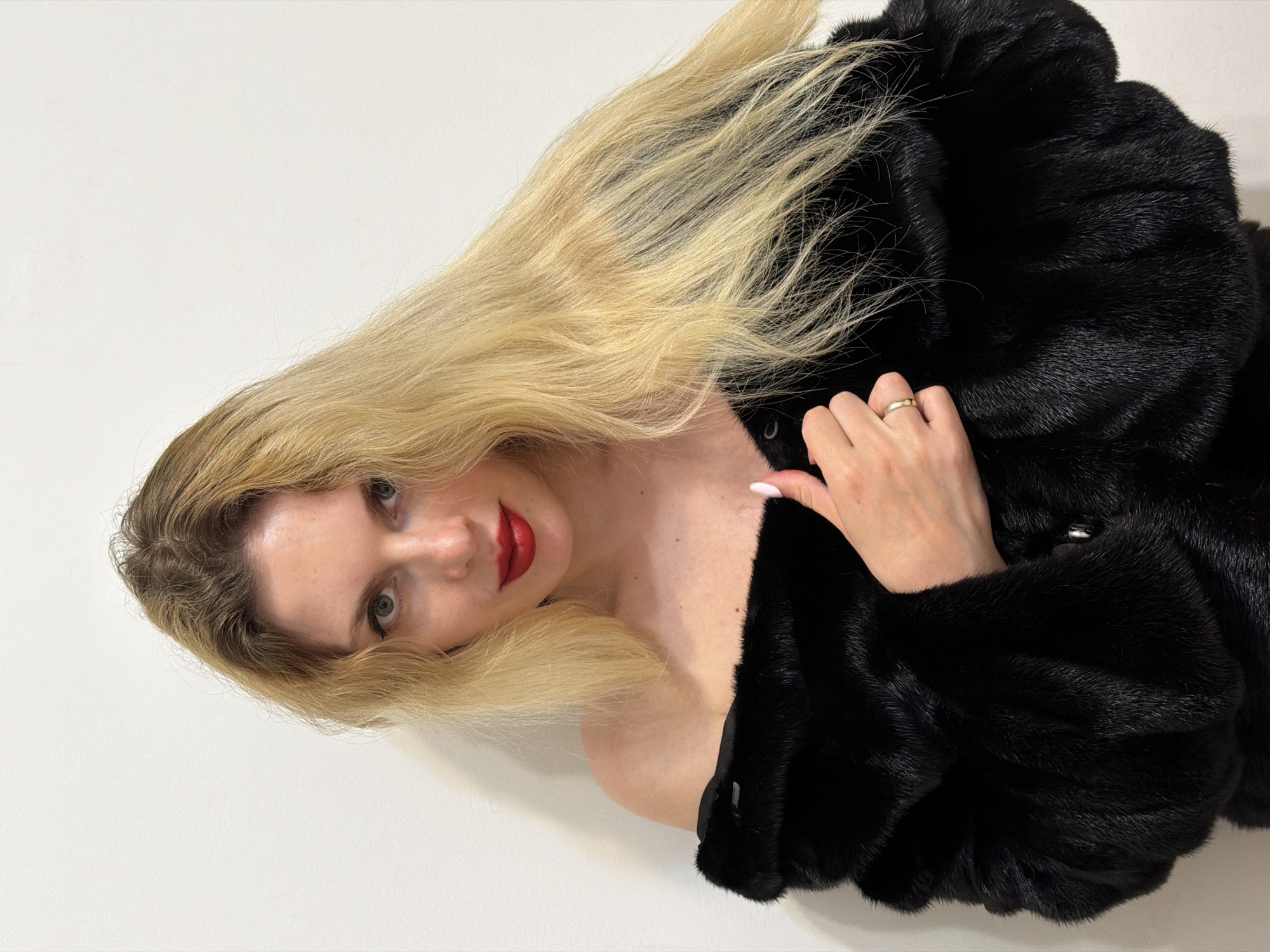 A fair-skinned woman with long, blonde hair is lying on a white surface, possibly a bed or floor. She is wearing bright red lipstick and a large, black fur coat, which she is holding closed with one hand. A simple gold ring is visible on her ring finger. The background is a plain white wall. There is no evidence of a location in Budva, Montenegro in this image.