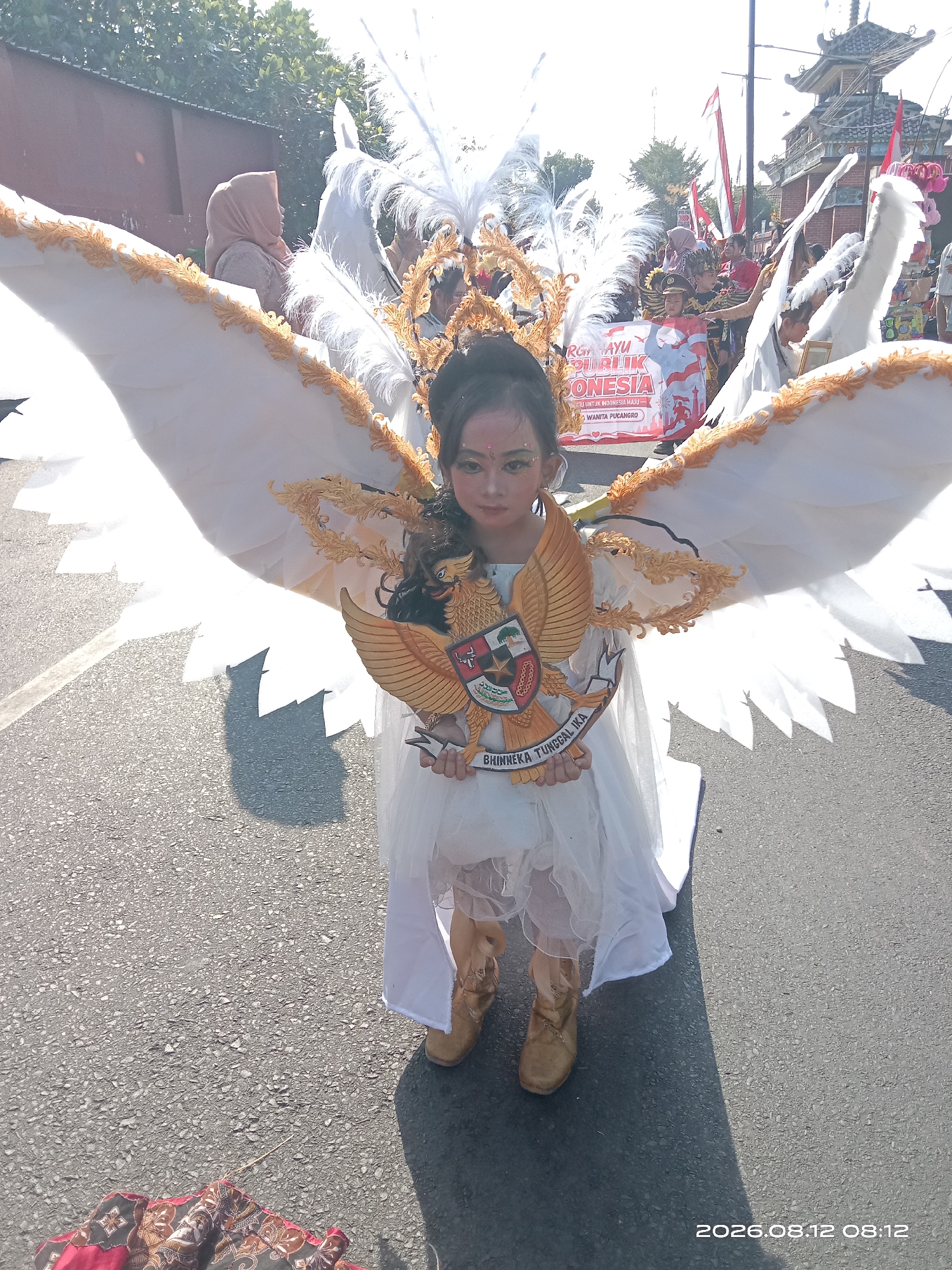 In Gudo, Indonesia, a young girl is dressed in a magnificent costume, likely for a parade or cultural event. She wears a white dress with ruffled layers, and large, elaborate white wings adorned with golden trim. On her chest, she holds a golden replica of the Garuda Pancasila, the national emblem of Indonesia, complete with its coat of arms and the motto "Bhinneka Tunggal Ika" (Unity in Diversity) inscribed at the bottom. Her face is painted with makeup, and her hair is dark and curly. The scene appears to be outdoors on a paved street, with a building and trees visible in the background. Other people, some holding banners, are also present, suggesting a public gathering. The sunlight casts a shadow on the ground. A timestamp in the lower right corner reads "2026.08.12 08:12".