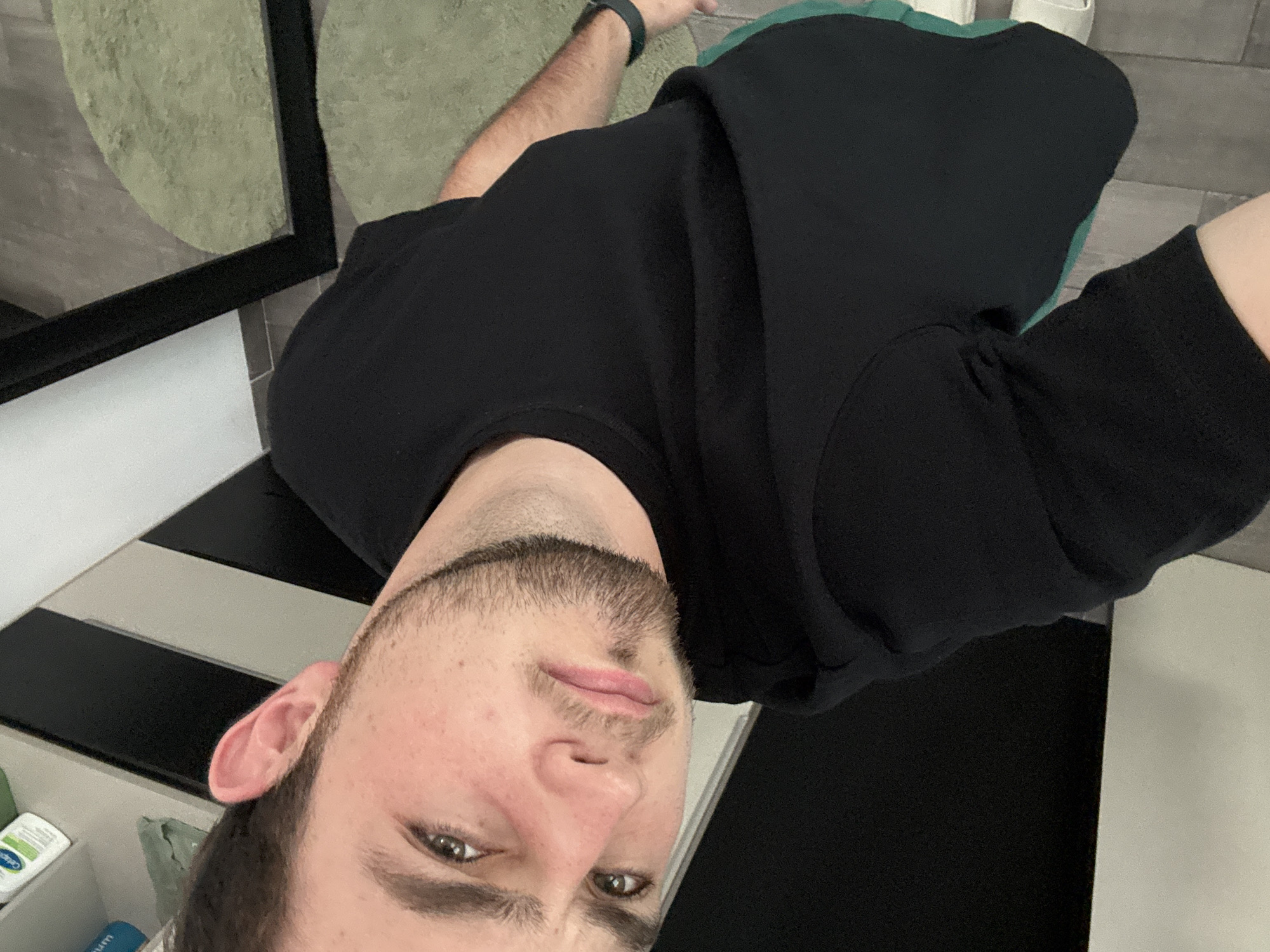 A man with a beard and short brown hair is angled towards the camera, wearing a black shirt. Behind him is a room with a black and white striped floor. There is a mirror on the left with a green rug behind it. Skincare products can be seen on a shelf in the lower left.
There is no indication in the image that it was taken in San Bernardino Tlaxcalancingo, Mexico.