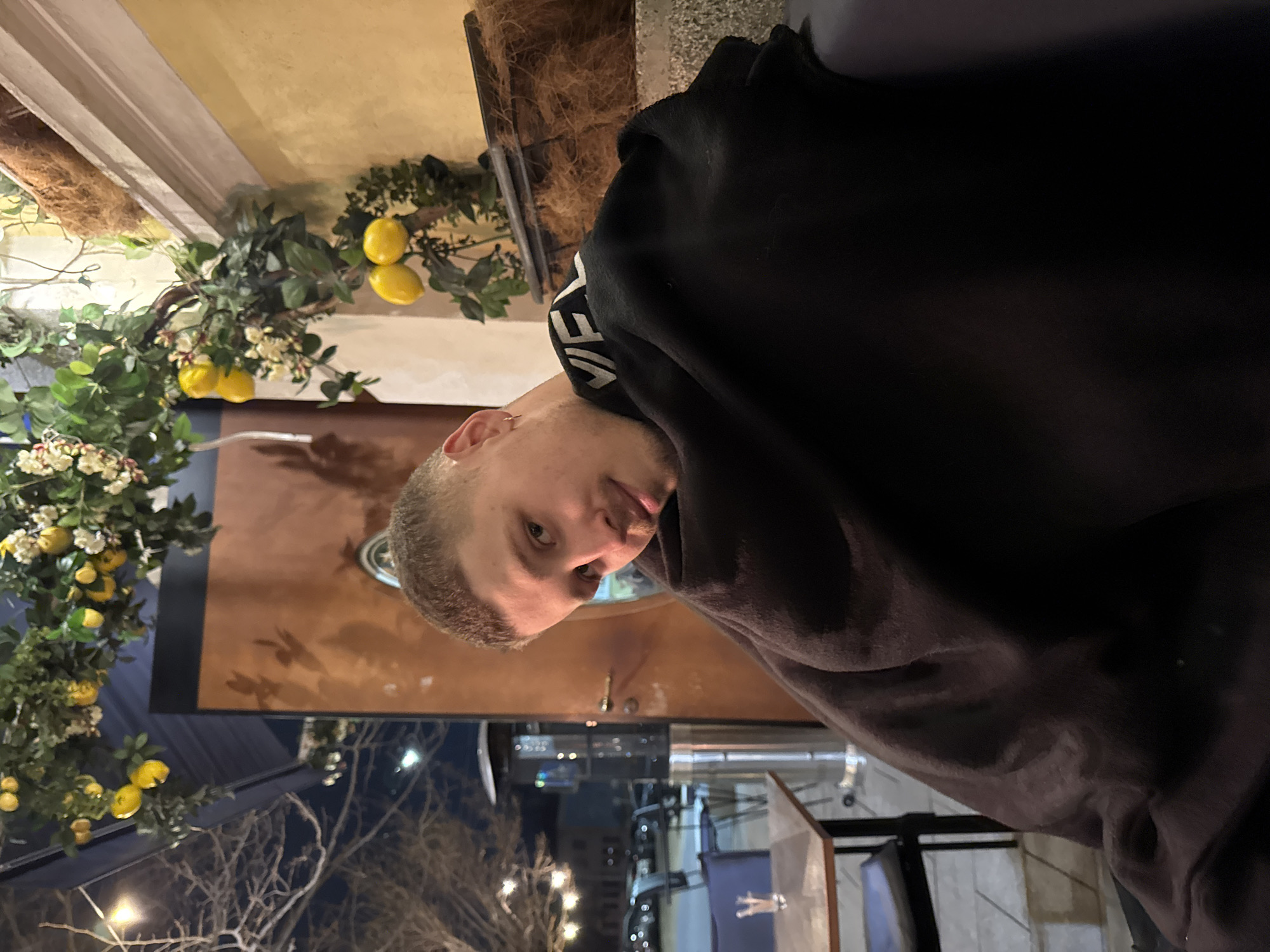 The scene depicts a male subject seated outdoors at night on what appears to be a restaurant or cafe patio in the city of Unknown, Unknown. The male, estimated to be in his late 20s to early 30s, has short dark hair and light facial stubble, with a small hoop earring in his left ear. He is wearing a dark garment, possibly a black sweatshirt or jacket, with the letters "VEL" visible in white on the collar/shoulder. A dark, possibly black, blanket or throw covers his lower torso. He is looking towards the camera with a slight smile.
The environment behind and around the subject includes artificial lemon branches with green leaves and white flowers draped overhead and along a light yellow wall to the subject's right. Below a white architectural ledge on the right wall, a planter containing dry fibrous material (resembling coconut coir) is visible. To the subject's left, a wooden-top table with a single blue chair holds a small clear glass containing decorative elements. Adjacent to the table, a tall, silver-colored patio heater is present. The background, visible to the left of the subject, shows bare tree branches, string lights, parked cars, and distant building lights, consistent with a nighttime outdoor setting. A warm brown wooden door or panel is located directly behind the subject.
