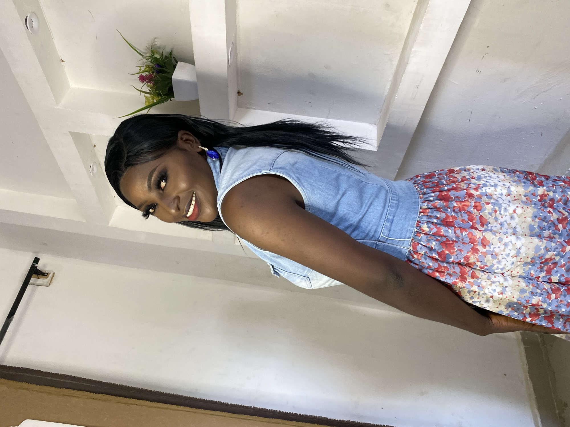 An African woman with dark skin and long black hair is posing for a portrait. She is wearing a sleeveless denim vest over a floral dress with a red, blue, and white pattern. Her makeup is noticeable with dark eyeliner and lipstick. She is also wearing a large blue earring. The woman is standing in front of a white wall with built-in shelves. On one of the shelves, there is a small potted plant with colorful flowers. The background appears to be indoors, possibly a studio or a room in a house, given the architectural details of the wall and the presence of a plant. The lighting suggests an indoor setting, possibly with natural light contributing to the overall brightness of the scene.