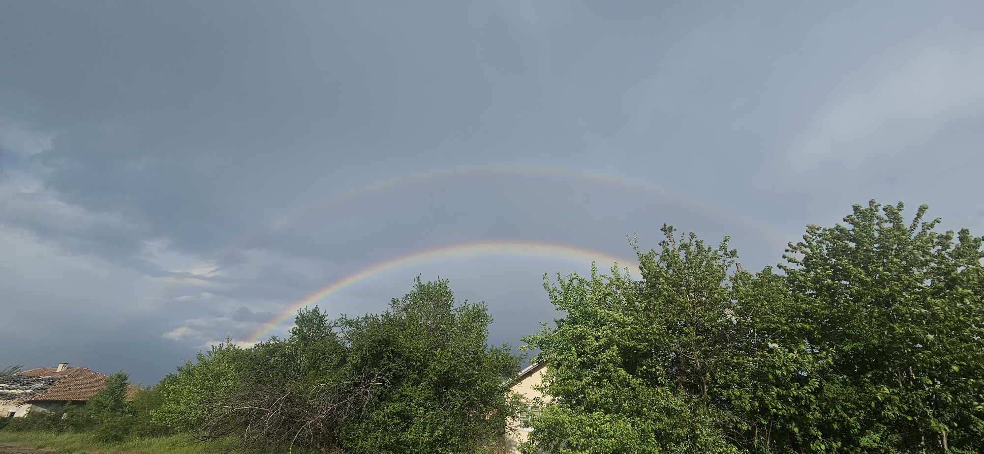 The image shows a double rainbow in the sky above Gutsal, Bulgaria. Green trees and a field occupy the foreground, partially obscuring two buildings. The building on the left has a damaged roof. The sky is overcast with dark clouds.