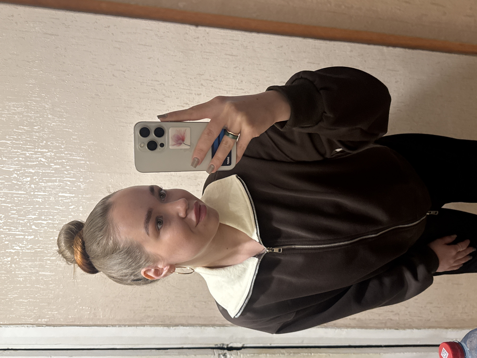 A young woman is taking a selfie indoors. She is holding a smartphone with a flower sticker on the back, revealing a ring on her finger and short, manicured nails. She is wearing a brown jacket with a white collar. Her hair is in a bun. The background shows a nondescript wall and a partial view of a window or ledge below. There is a water bottle with a red cap visible in the lower right corner. There is no verifiable information in the image to confirm the location as the city of Chaikovsky, Russia.