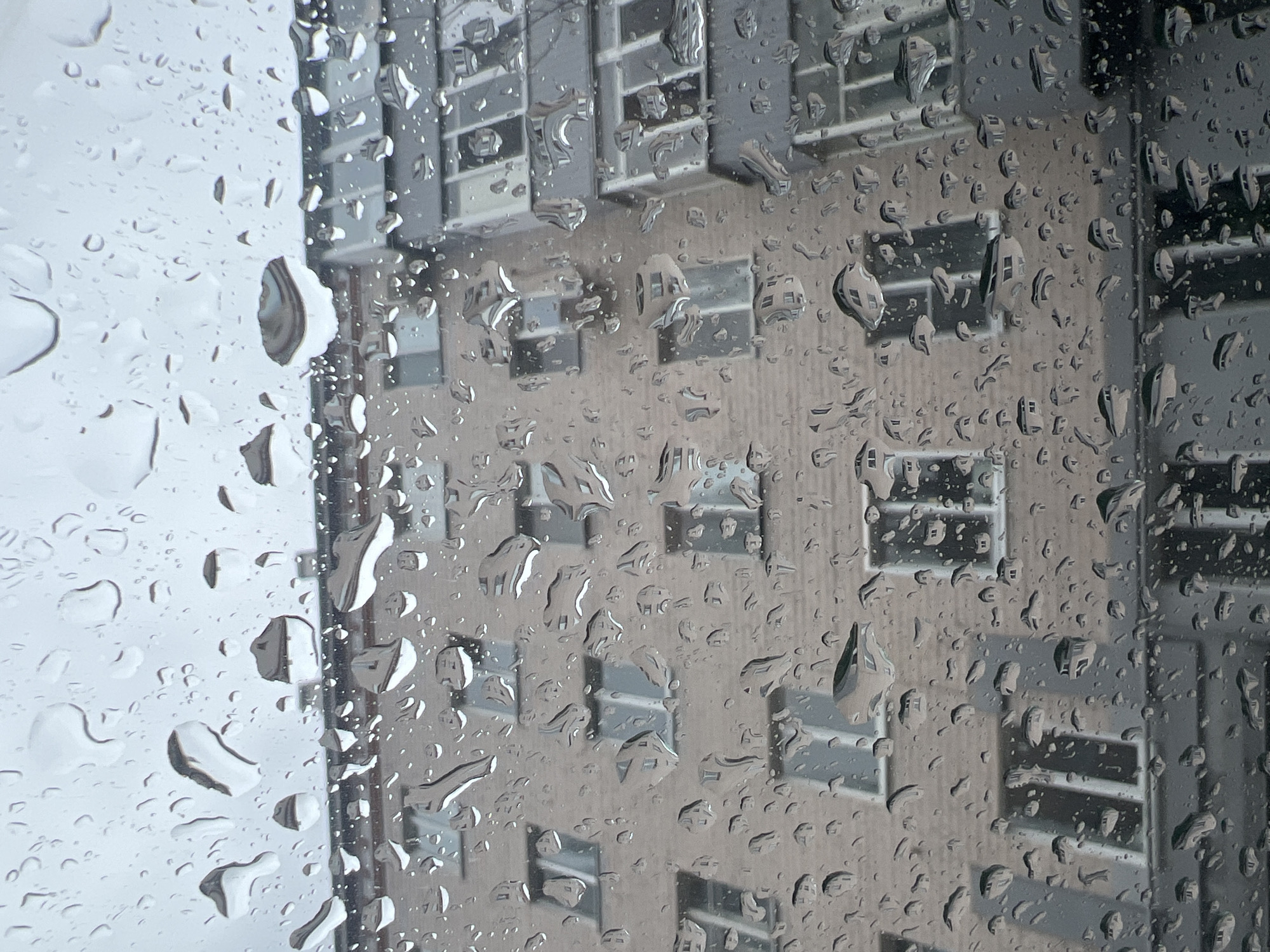 The image shows a view from inside, looking through a window covered in raindrops. The blurred reflection of a tall building with many windows is visible through the rain. The building appears to have a light-colored facade with dark window frames. The raindrops on the window obscure the details of the building, creating a distorted and indistinct view. The location is Kazan, Russia.