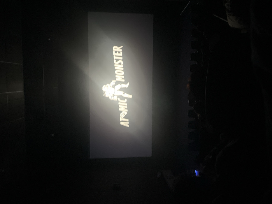 A dark, interior view of a movie theater in Mexico City, Mexico, displays a large projection screen centered with the title "ATOMIC MONSTER." The text is rendered in a light, stylized font, with an atomic symbol replacing the 'O' in "ATOMIC." To the right of the title, a light-colored, bipedal figure in a bulky suit or armor with a helmet and backpack is depicted. Below the screen, the silhouettes of multiple audience members occupy rows of dark theater seats. One individual on the left holds a mobile device, which emits a distinct blue light illuminating their hand and immediate area. The surrounding environment of the cinema hall, including walls and ceiling, is uniformly dark, consistent with a viewing environment.