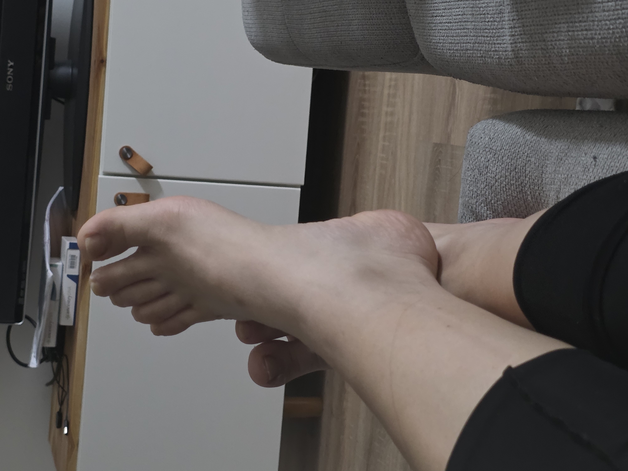 An overhead shot captures a pair of bare feet crossed over each other, resting on a gray upholstered piece of furniture, likely a sofa. The feet are fair-skinned, with visible toes. One foot is positioned slightly above the other. Dark-colored fabric, possibly leggings or shorts, is visible around the ankles and lower legs. The background reveals a wooden floor and a white cabinet with two brown, leather drawer pulls. Above the cabinet, a portion of a black television screen with a "SONY" logo is visible, along with some papers and cables on a wooden shelf. The overall scene suggests an indoor setting, possibly a living room.