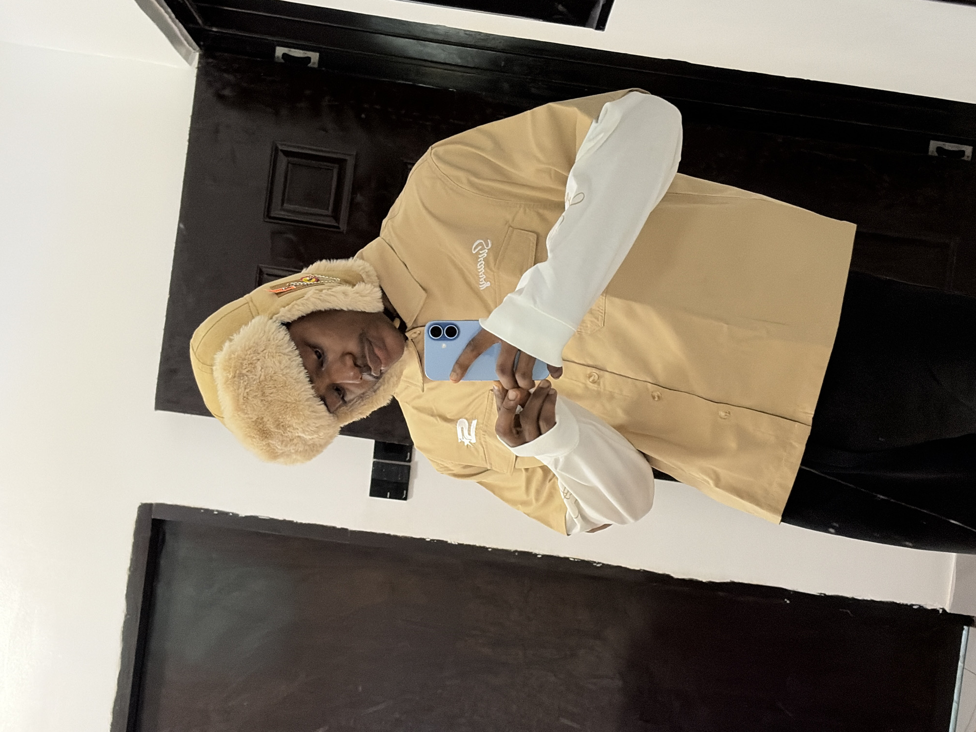 A young Black male, wearing a tan collared shirt over a white long-sleeved shirt, black pants, and a tan trapper hat with fur trim, stands indoors and takes a selfie with a light blue smartphone. The male is looking down at his phone. He has a mustache and is holding the phone in his left hand, with his right hand near the phone. The background consists of a white wall with two dark doors. The image is likely taken in Igboefon, Nigeria, as indicated by the provided location.