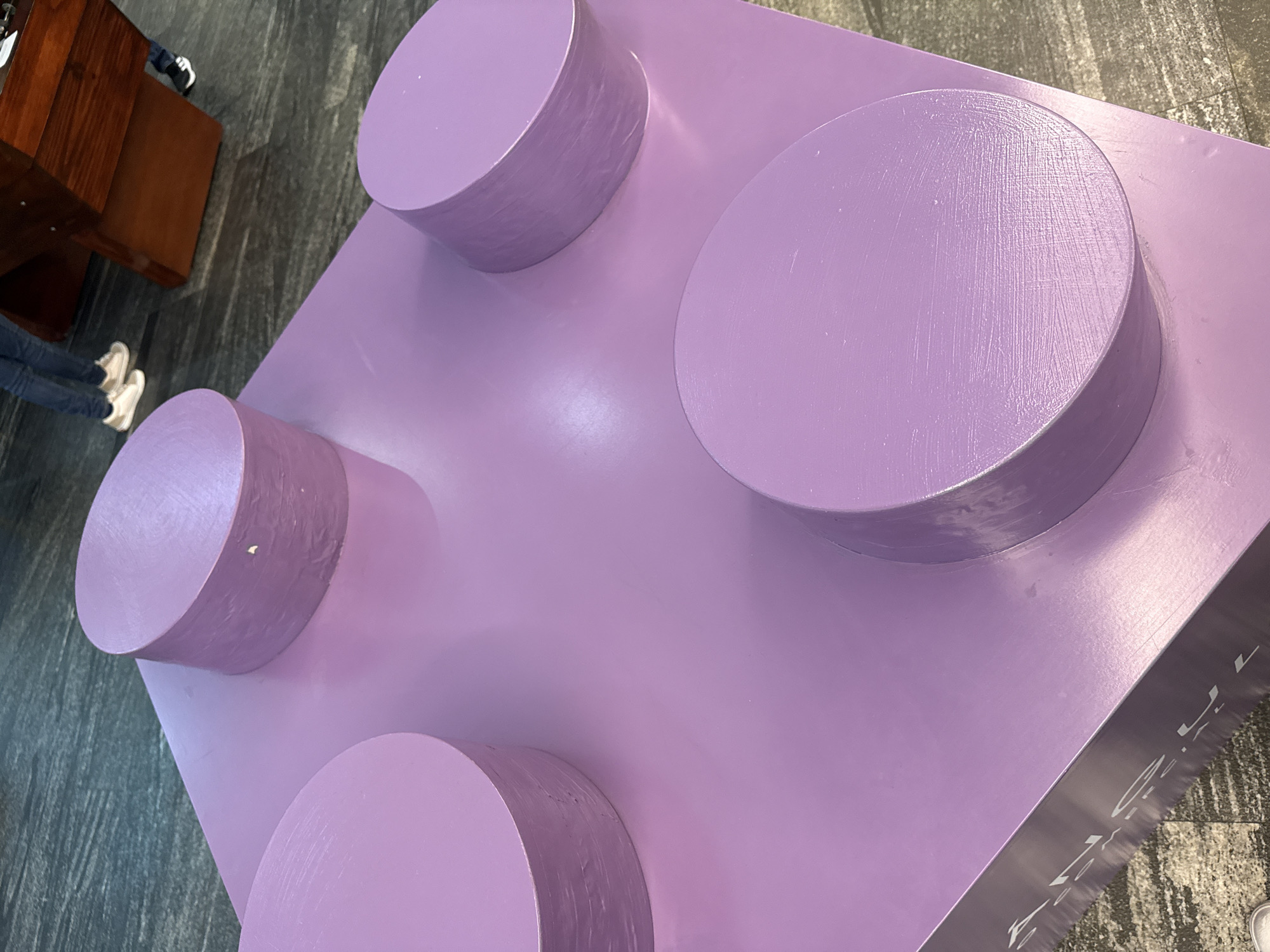 A top-down view shows an oversized, four-studded lavender-colored Lego brick. Partial views of a wooden desk and a person's legs are visible to the left of the brick. The location is the city of Puebla, Mexico.