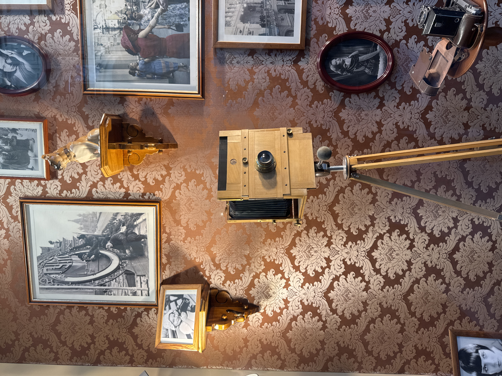 The image depicts a wall covered in a patterned brown damask wallpaper. Several framed photographs and a vintage camera are displayed on the wall and a nearby tripod.
Prominently featured is a large, framed black and white photograph of several people working on what appears to be a clock face or large mechanical device, with a cityscape in the background. To the left of this, a smaller framed photograph shows a child holding a camera.
Below these, a wooden shelf holds a ceramic figurine of a dog. To the right, a large, wooden bellows camera on a tripod is positioned centrally. Further to the right, a framed photograph in an oval red frame shows a woman posing. Below that, another framed photograph shows a historical cityscape.
On a small table in the lower right, a vintage camera bag and a second vintage camera are visible. In the lower left corner, a framed photograph of a woman with dark bobbed hair is partially visible.
The overall impression is a curated display of vintage photography equipment and historical images. Based on the architectural style visible in the large photograph on the wall, it is plausible that the setting depicted in that photograph is Moscow, Russia, possibly Red Square, given the presence of onion domes in the background. However, the exact location of the displayed items is not specified within the image itself.