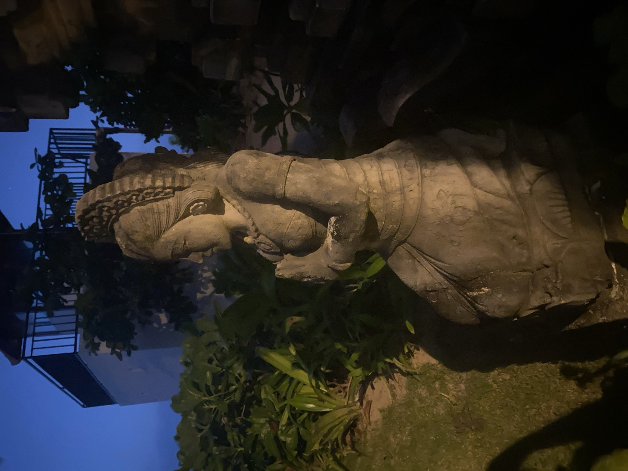 The image depicts a stone statue lying on its side in Kuta Selatan, Indonesia. The statue is weathered and appears to be of a human figure with decorative elements. It is surrounded by vegetation, including green plants and trees. In the background, a building with a metal balcony is visible against a dark blue twilight sky. The scene is dimly lit.