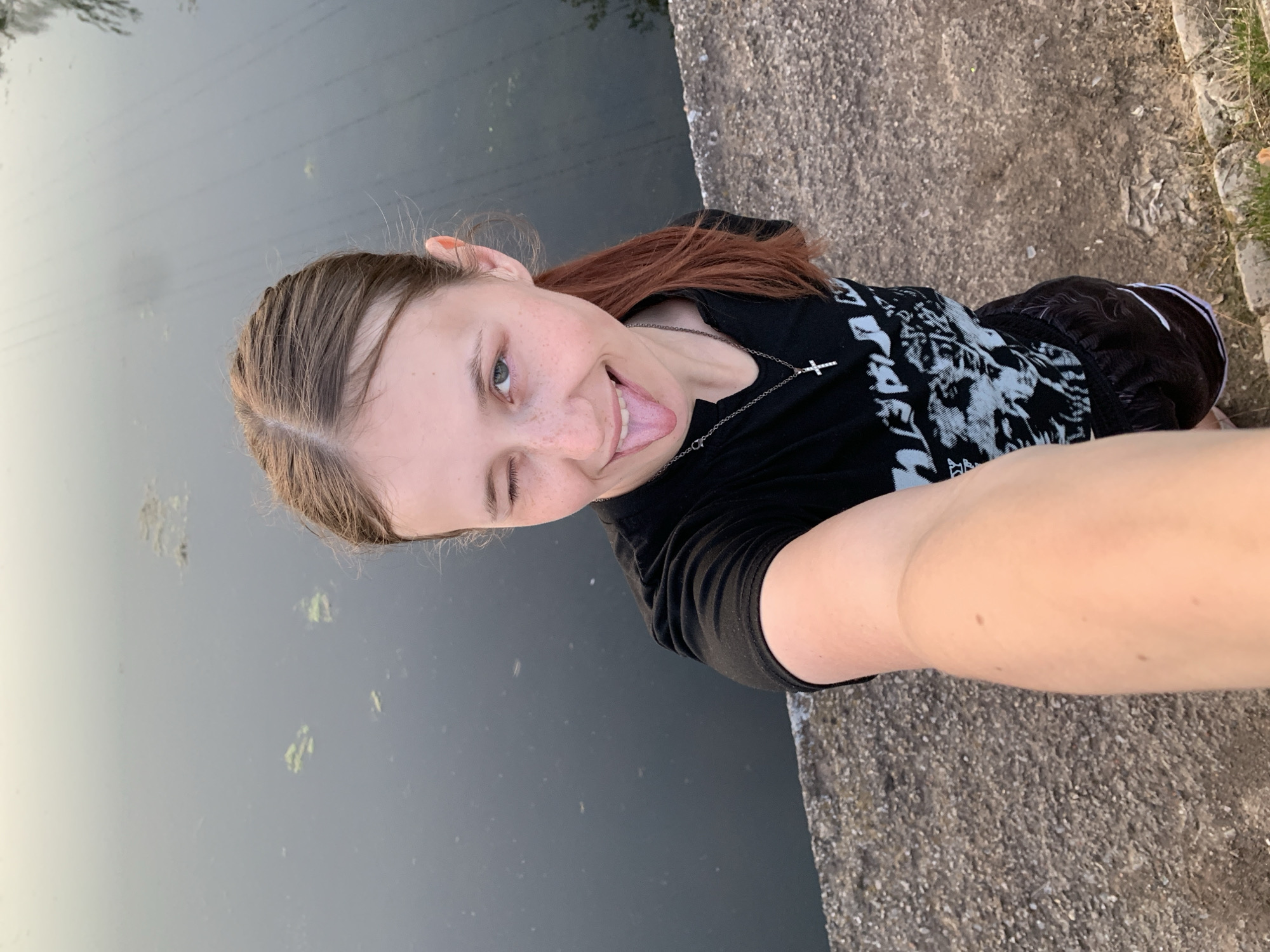 A person with light skin and light brown hair with reddish highlights stands on a concrete structure next to a body of water. The person is winking and sticking out their tongue, taking a selfie. They are wearing a black t-shirt with white graphics and black shorts with white stripes. A silver necklace with a cross pendant is visible. The water has some green algae or debris floating on the surface. The concrete structure appears to be a bridge or retaining wall. Based on the description provided, the city is Фоки, Russia.