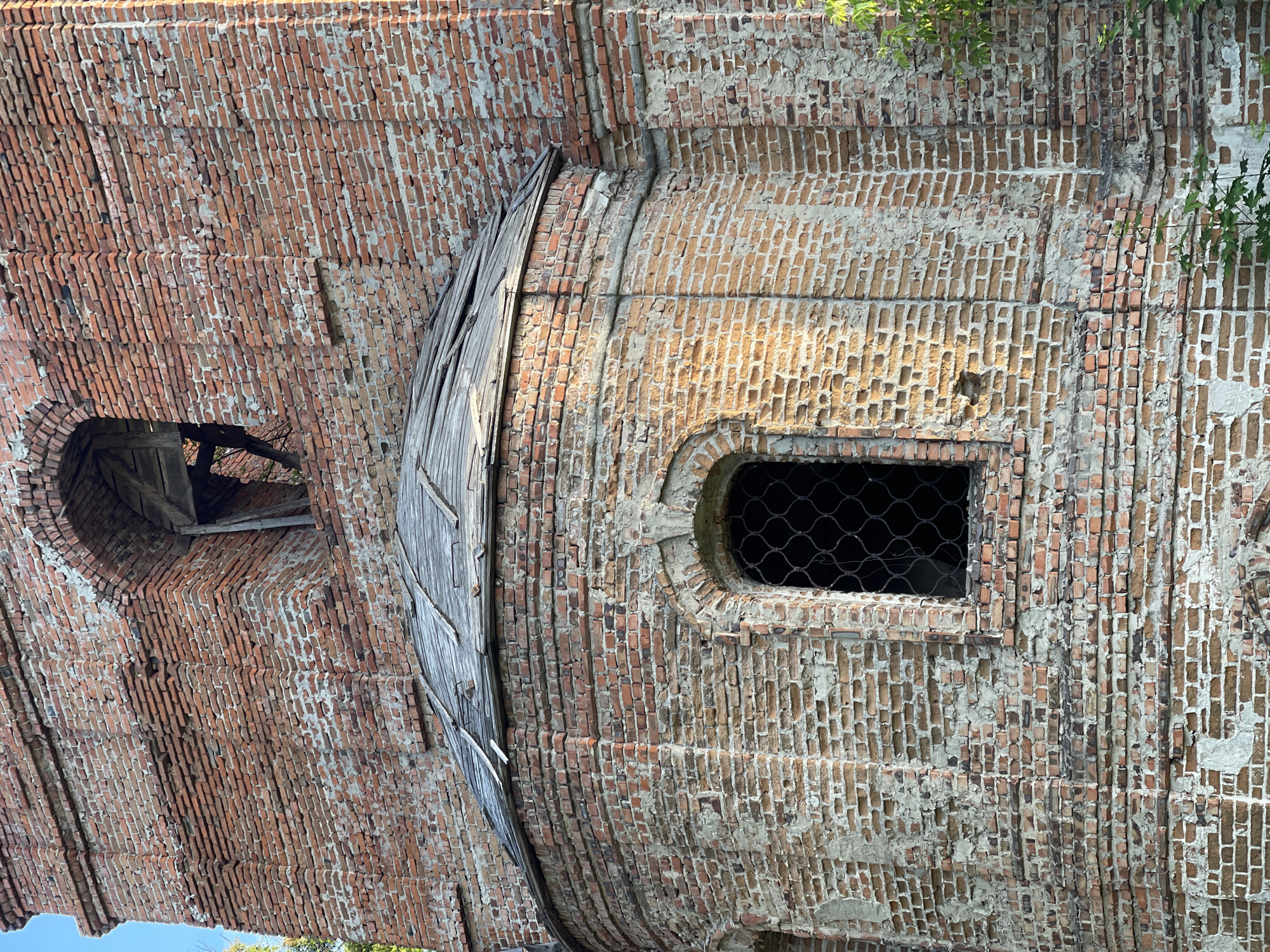 A close-up, full-frame, eye-level, vertical shot shows the weathered, red brick facade of an old building, likely a church or historical structure, bathed in natural daylight. The bricks are unevenly laid, with visible mortar that has deteriorated in many areas, revealing patches of lighter plaster or concrete underneath. The wall displays architectural features such as arched window openings and a curved section topped with a dilapidated wooden roof composed of decaying planks. One arched window is partially visible, covered by a dark, lattice-like metal grate. Above this, another arched opening in the upper brickwork shows exposed wooden beams and structural elements. Small patches of green foliage are present at the bottom and right edges of the frame, suggesting the building is surrounded by vegetation. The lighting creates shadows that highlight the texture and imperfections of the aged brickwork.