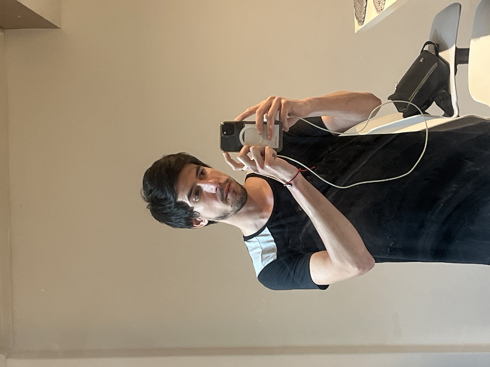 A selfie depicts a man with dark hair and a trimmed beard. He is wearing a black t-shirt with a white stripe on the left sleeve, holding a smartphone up to take the picture. The background is a plain wall. A black bag hangs on a white chair in the upper right corner. The location of the photo is unknown.