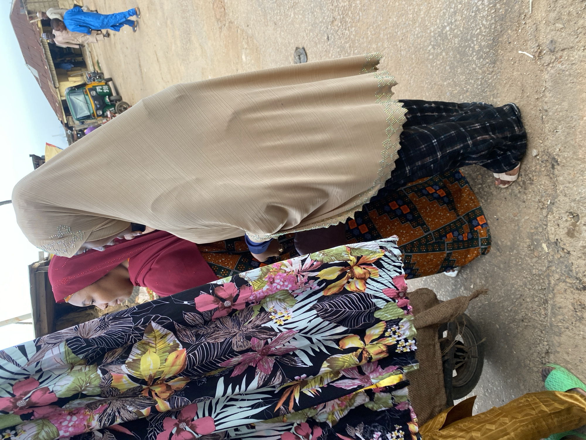 In Jos, Nigeria, two women are standing in a market. One woman, on the left, wears a red hijab and a patterned orange and black dress. She is looking to her left at a large piece of fabric with a floral print. The second woman, on the right, wears a beige hijab and a black and white plaid skirt. Her right hand is visible, with a ring on her finger. Behind them, a small green and yellow motorized tricycle, often called an "okada" or "keke napep" in Nigeria, is parked. A young boy in a blue outfit is walking past the tricycle. The ground is dirt and gravel, and buildings with corrugated metal roofs are visible in the background. The sky is overcast.