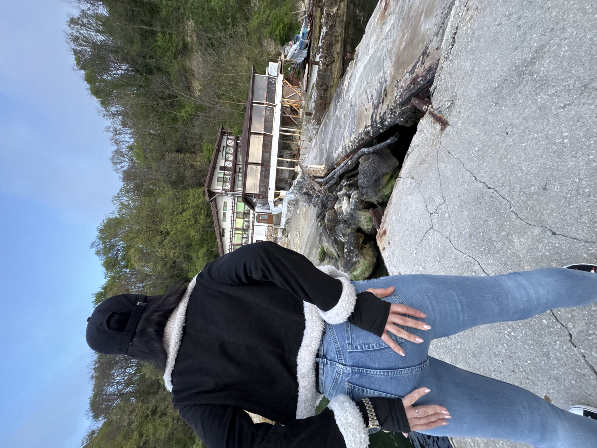 A medium shot of an individual with long, dark hair wearing a black baseball cap, a black jacket with white fur trim, and blue jeans, posed on a concrete surface. The individual is positioned with their back to the viewer, hands placed on their rear, and wearing a watch on one wrist.
The background includes a two-story building with a dark roof and white exterior, situated near a concrete embankment. Vegetation covers a hillside behind the building, with the sky partially visible. The setting appears to be a coastal area, with features resembling the city of Varna, Bulgaria. Evidence of erosion is visible on the concrete, and a rocky area borders the edge.