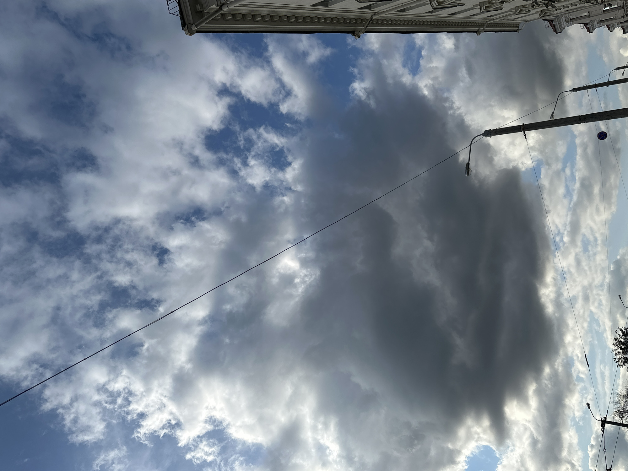 The image depicts a dramatic sky with large, dark storm clouds dominating the upper portion, interspersed with patches of bright blue sky and smaller, white clouds. A thin black wire stretches diagonally across the frame from the top left to the bottom right. In the foreground, to the right, a portion of a white building with classical architectural details, including cornices and windows, is visible. Several lampposts with lights are present, with one prominent lamppost in the center, and others further back. The lower part of the image shows trees and more wires against a brighter, lighter sky with scattered clouds. The scene appears to be an urban environment, likely on a street, given the presence of buildings, lampposts, and wires. The overall impression is of an impending or passing storm.