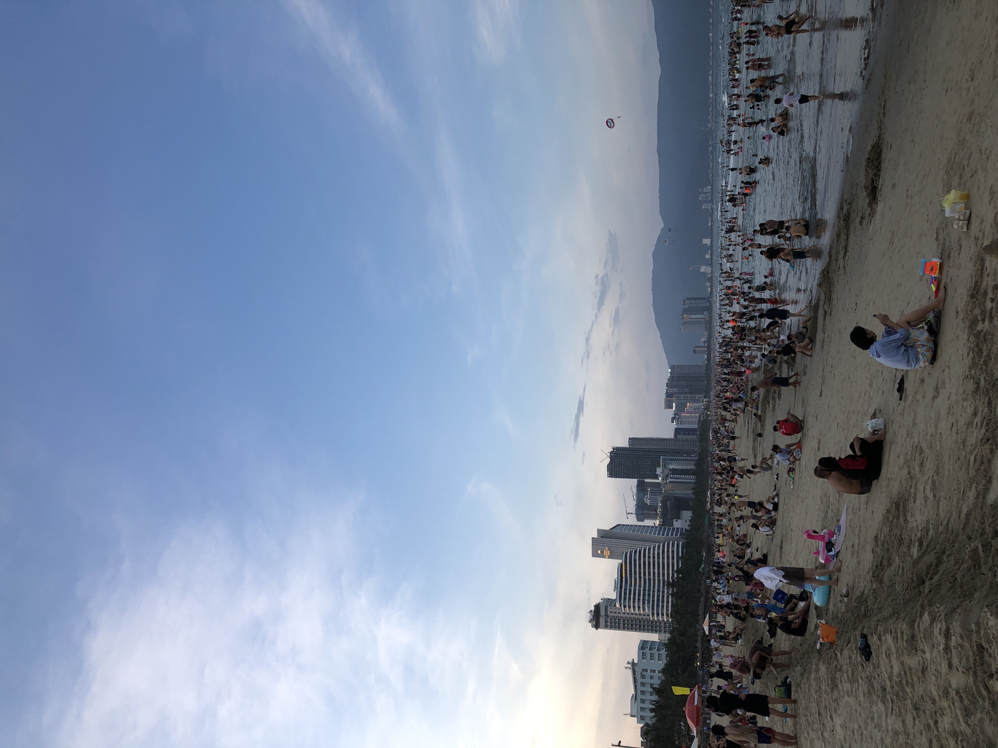 This media file captures a bustling beach scene in Đà Nẵng, Vietnam, likely during the late afternoon or early evening. The sky is a clear blue with wispy clouds, suggesting fair weather. A large crowd of people are enjoying the beach, with many wading in the shallow water or walking along the shoreline. The ocean appears calm, reflecting the sky.
In the background, a modern cityscape rises, characterized by numerous tall buildings and high-rises, indicating an urban coastal location. The buildings suggest a developed tourist area. Further in the distance, a hazy mountain range borders the cityscape.
Several recreational activities are visible. One prominent feature is a parasailer in the sky, a common beach attraction. There are also smaller parachutes or kites further out in the water. On the sandy beach, people are seated, lying down, or playing, with some children's toys visible. The overall atmosphere appears relaxed and communal, with many individuals and groups enjoying leisure time. The lighting suggests it is not midday, likely transitioning towards sunset. No visible text is present in the image.