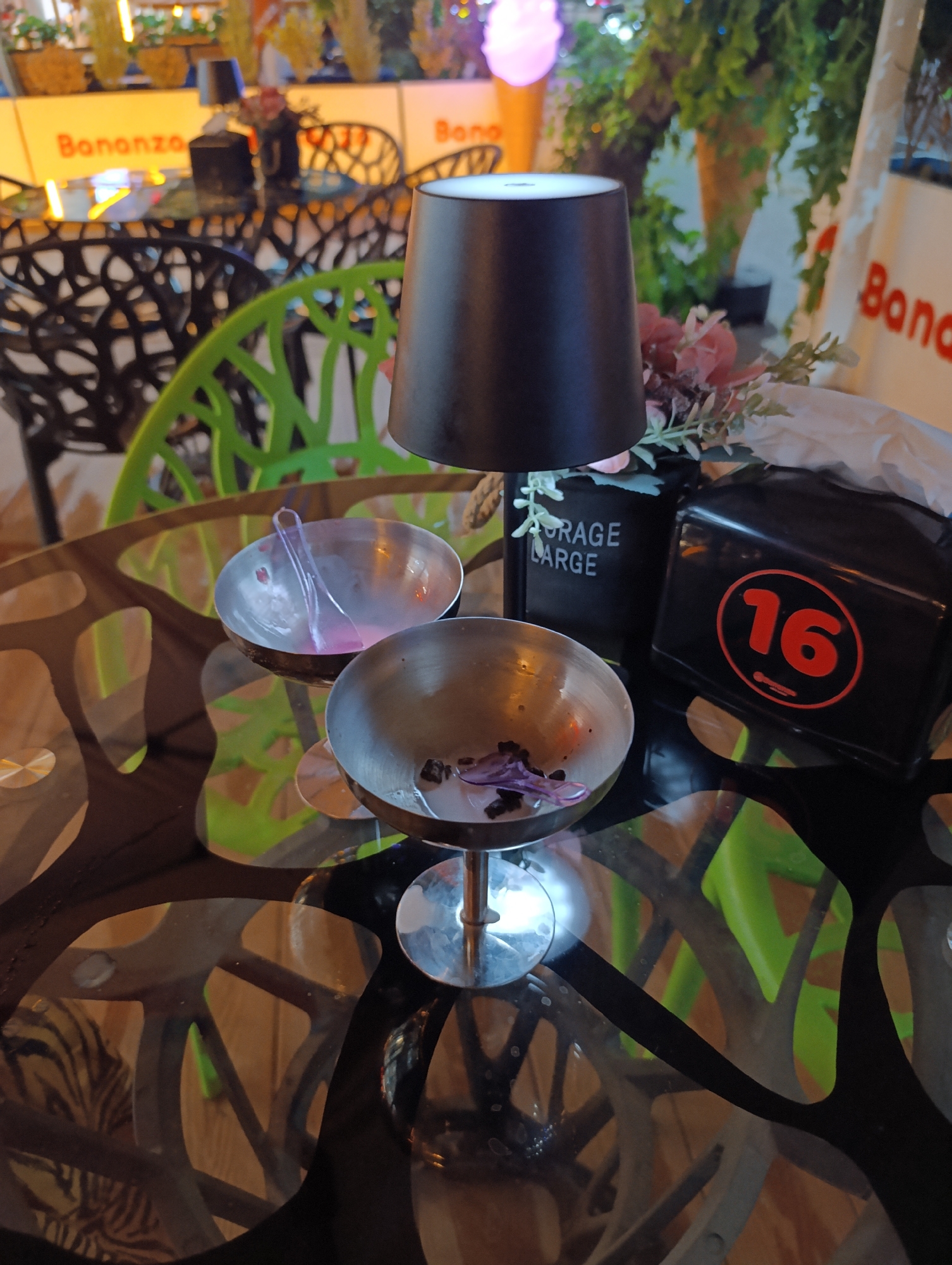 This is a close-up, eye-level shot of a glass-topped table at what appears to be an outdoor cafe or restaurant. Two metal bowls, one with a pink residue and a plastic spoon, and another with some dark remnants, sit on the table, suggesting a recently finished meal or dessert. To the right of the bowls is a black napkin dispenser with the number "16" in a red circle. Behind it, a black container with the words "STORAGE" and "LARGE" printed on it holds a small arrangement of artificial flowers. A black lamp with a conical shade is positioned between the flowers and the bowls, its light illuminating the table.
The background is softly blurred, showing tables and chairs in an outdoor setting. Some chairs are black and intricately designed, while others are bright green and plastic. In the distance, a sign with red lettering that reads "Bonanza" is visible. The overall ambiance suggests it is evening or night, given the artificial lighting and the mood it creates. There are no people visible in the frame. The image focuses on the table setting, giving a sense of a quiet, perhaps solitary, moment after a meal.