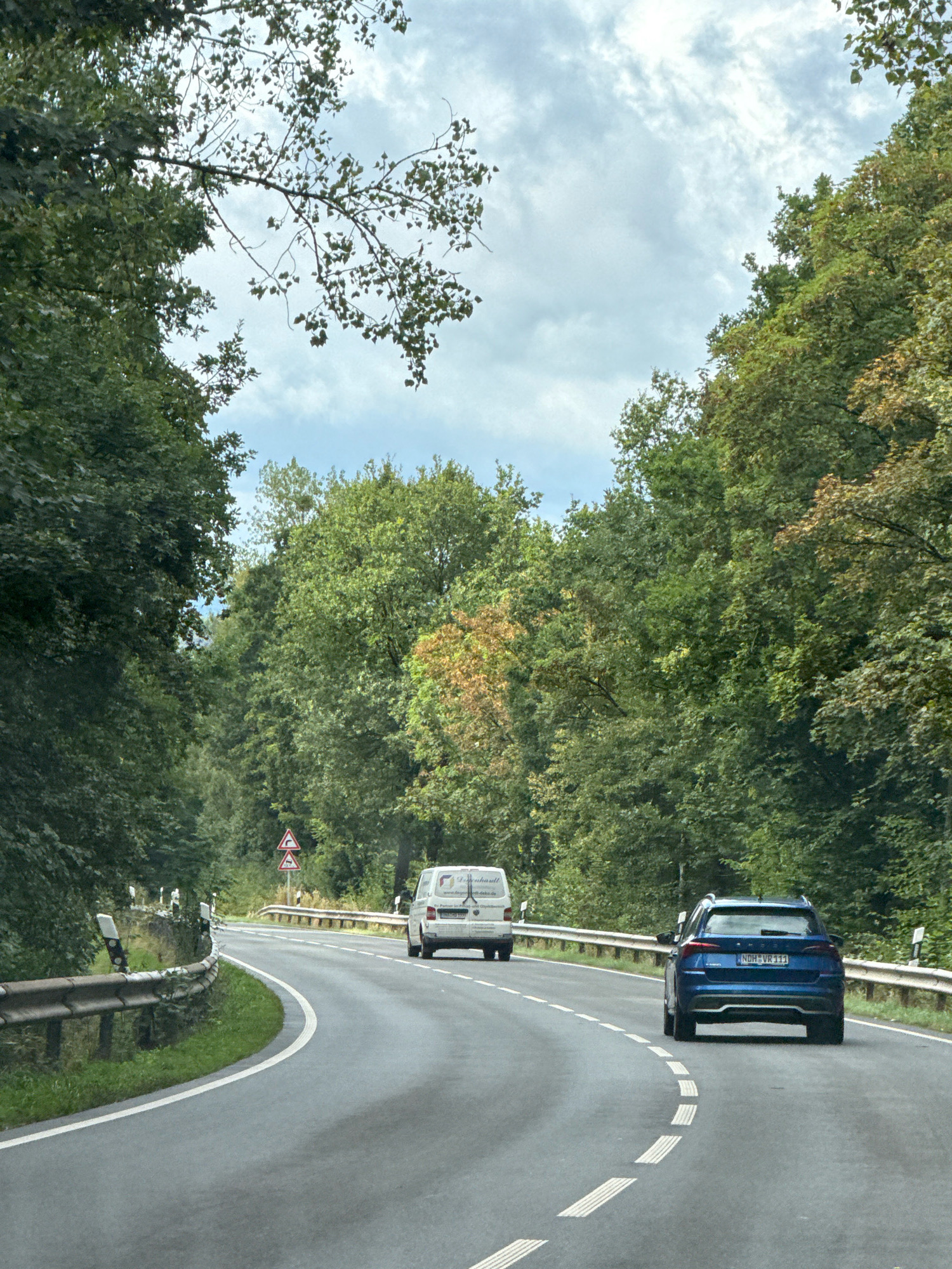 The image depicts a two-lane road in a wooded area, likely within or near the town of Bad Sachsa, Germany. The road curves to the right, bordered by a metal guardrail on both sides. Dense green trees line the road, with some showing hints of autumn foliage. A blue SUV with license plate "NDH VR 111" is driving in the right lane, ahead of the viewer. In the left lane, further ahead, is a white van with text and a logo visible on its side, suggesting it is a commercial vehicle. Two triangular road signs with red borders are posted on the left side of the road, indicating a curve ahead. The sky is overcast with scattered clouds. The scene appears to be captured from inside another vehicle, moving along the road.
