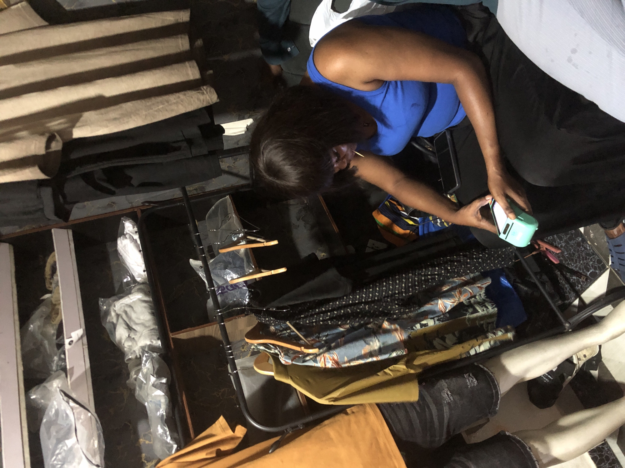 The image depicts an indoor scene, possibly a room or a shop, with clothing items displayed and stored. In the foreground, a person wearing a blue tank top and black pants is seated and holding a small, light blue electronic device with white accents. This device appears to be a portable card reader or payment terminal. Their left hand is holding the device, and their right hand is near it, suggesting an interaction. A dark smartphone is placed on their lap.
Behind the person, clothing is visible on hangers, including a yellow shirt, a patterned shirt with a floral design, and a black patterned garment. Further back, shelves are laden with clothing items, some of which are packaged in plastic. A mannequin dressed in a yellow shirt and denim shorts stands to the left of the frame. The overall environment suggests a retail or storage space for clothing. The image is taken from a medium shot, focusing on the person and their immediate surroundings. The lighting is dim, casting shadows. The location in the city of Ado, Nigeria, is not explicitly evident from visual cues within the image itself.