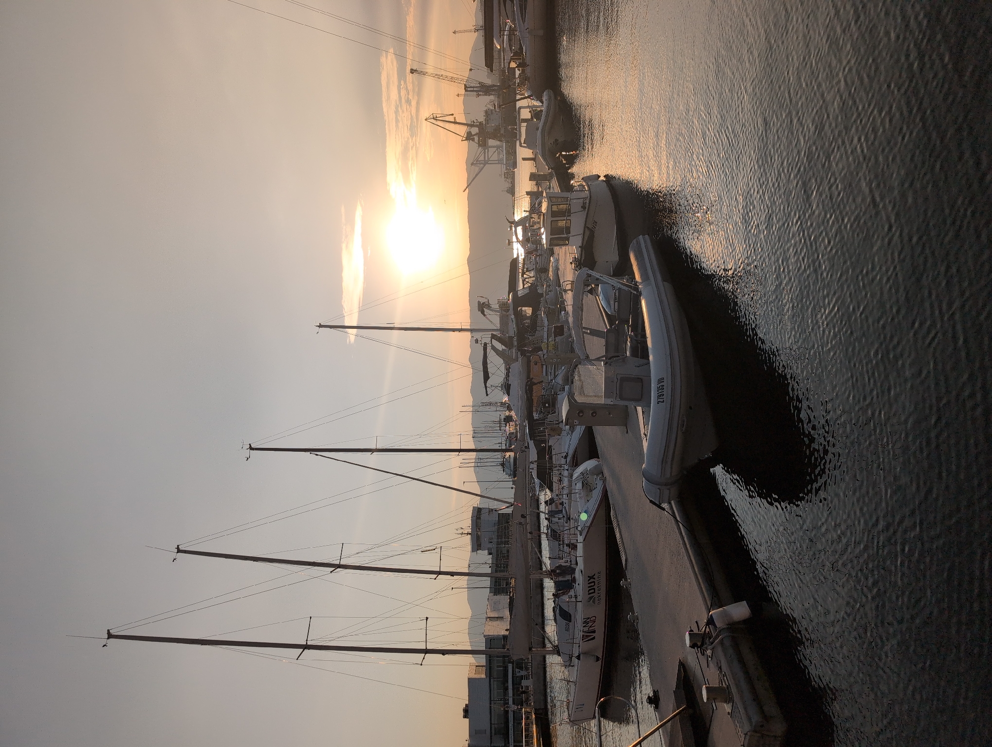 The image depicts a harbor scene at sunset in the city of Rijeka, Croatia. In the foreground, several boats are moored to a dock. Among them is a white inflatable dinghy with the registration number "279135 VD". Further back, a white sailboat with "DUX nekretnine" branding is visible, along with other sailboats and smaller motorboats. In the background, a port area with cranes and industrial buildings is silhouetted against the setting sun. The sky is a soft gradient of orange, pink, and pale blue, with the sun casting a warm glow on the water. The overall impression is one of a tranquil evening in a bustling port city.