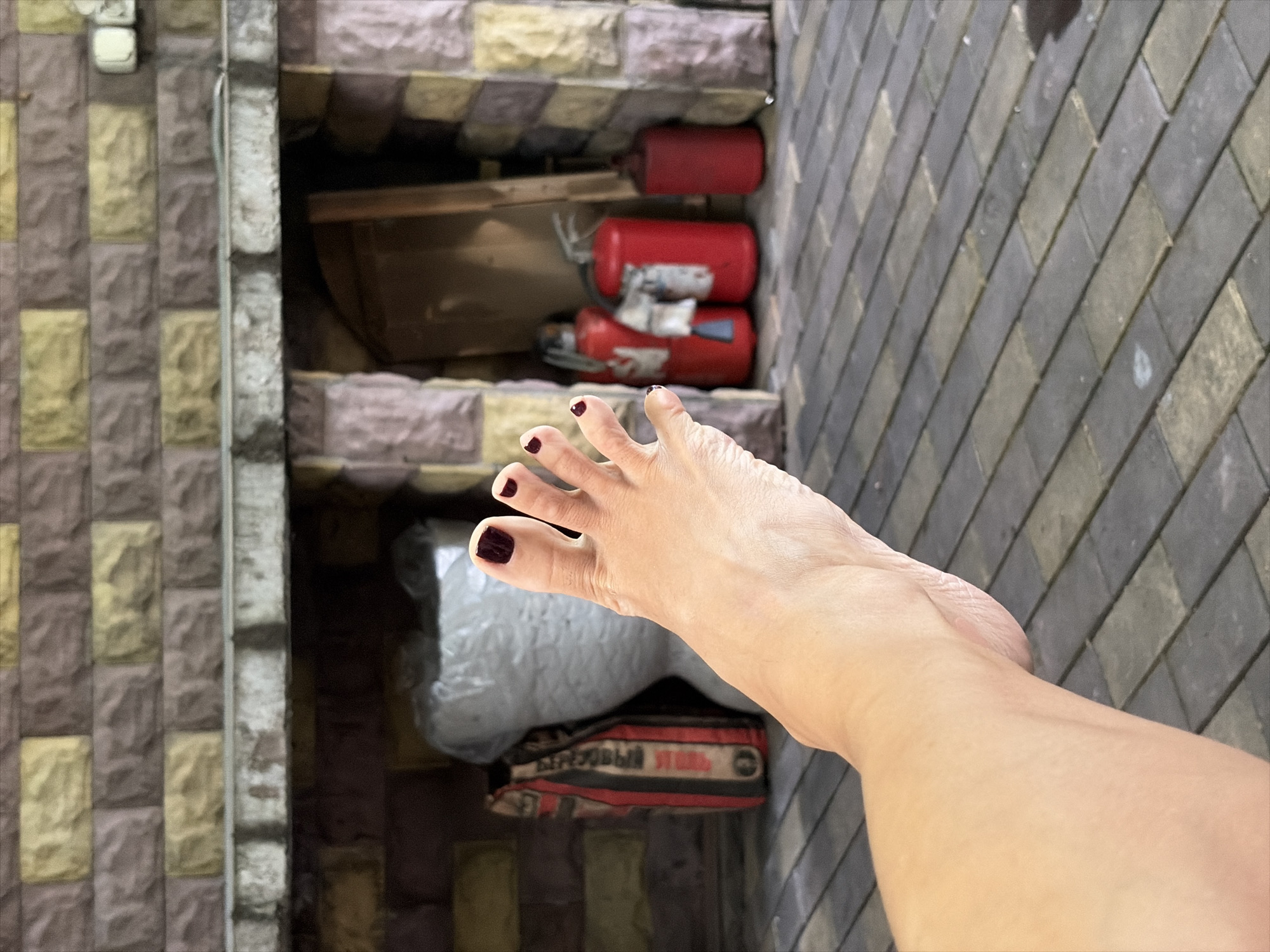 A human foot with dark nail polish is positioned in the foreground, extending from the left side of the frame. The foot is bare and appears to be resting on a grey and yellow brick patio. In the background, there is a stone structure resembling a fireplace or an outdoor oven, with several red fire extinguishers and what appears to be a bag of cement or similar material within its alcoves. The overall setting suggests an outdoor, possibly domestic or industrial, area in Balashikha, Russia.