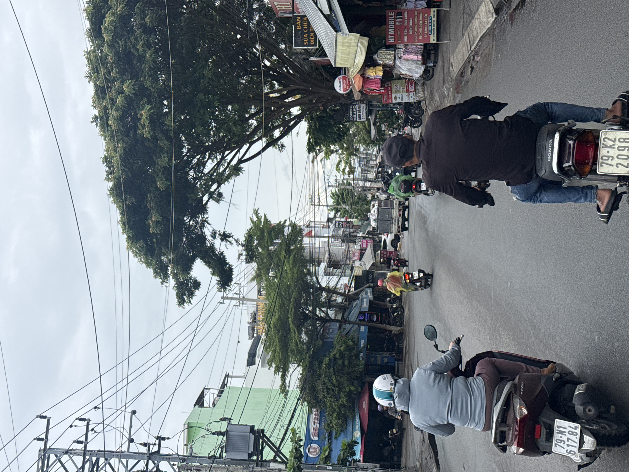 A street view in Nha Trang, Vietnam, shows a cloudy day with a road and buildings. In the foreground, two individuals are riding scooters, one in a grey hoodie and burgundy pants, the other in a dark shirt and blue jeans. Both are facing away from the camera. In the background, other people on scooters and a truck are visible. Businesses line the street, displaying signs in Vietnamese. Power lines crisscross above the street, indicating a dense urban environment. Trees provide some shade along the road. The overall impression is a typical street scene in a bustling Vietnamese city.