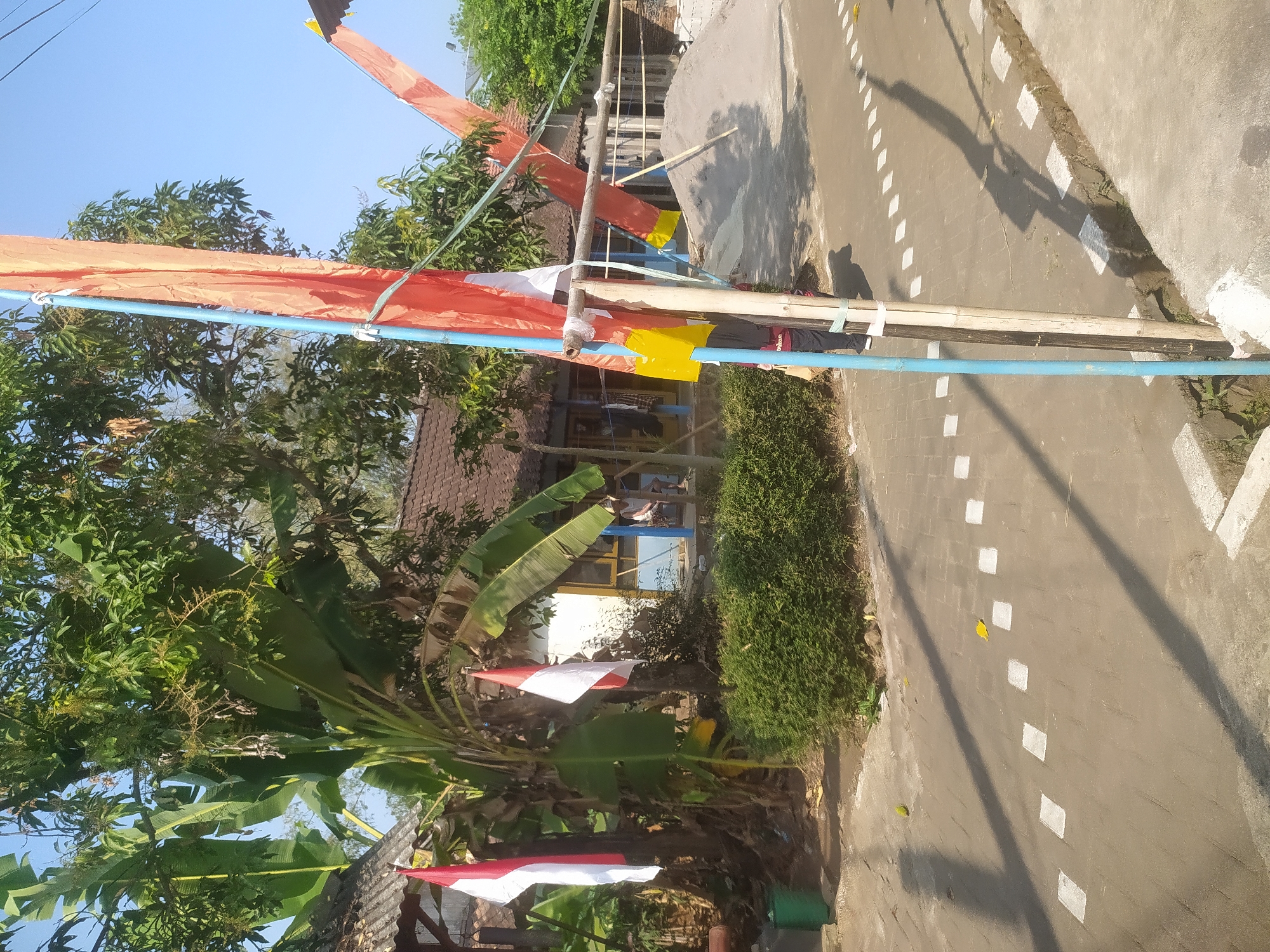 The image depicts a street scene in Lopait, Indonesia. The foreground shows a paved road with white dashed markings. To the right, a drainage ditch runs along the side of the road. In the mid-ground, a pile of sand is visible, with some construction materials nearby. Two Indonesian flags, red and white, are mounted on poles amidst lush green vegetation, including banana trees. Orange and yellow banners are also displayed on poles. Behind the vegetation, traditional Indonesian houses with tiled roofs can be seen. The sky is clear and blue. The image appears to be taken during the day, with sunlight casting shadows on the road. A person is partially visible in the mid-ground, walking towards the right.