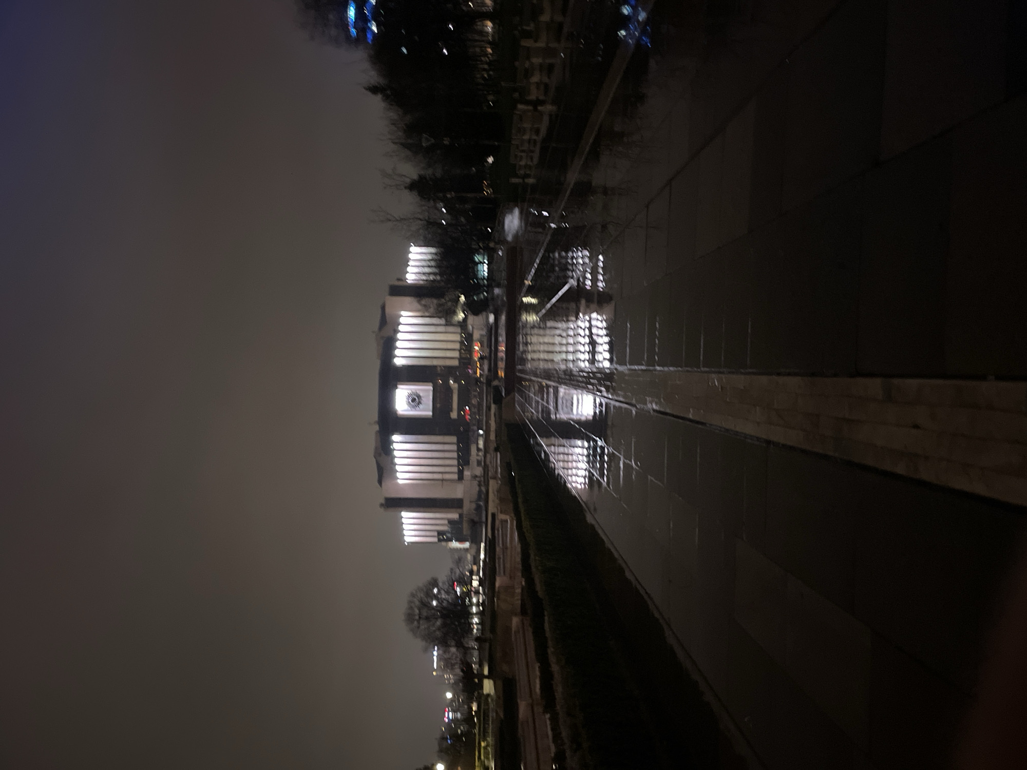 The image shows a night scene in Sofia, Bulgaria, likely near the Palace of Justice. The building is lit up and its facade is visible, with a large clock face. A wet, reflective surface, possibly a paved area or path, is in the foreground, reflecting the lights of the building. The surrounding environment is dark, with trees and streetlights adding to the cityscape. The sky is overcast. There are no people visible in the immediate scene.