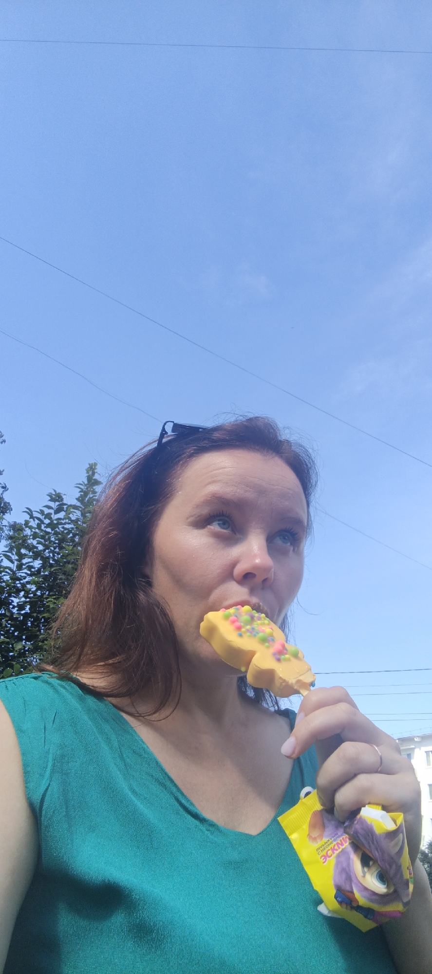 An overhead, medium shot shows a woman eating a yellow, bear-shaped popsicle with sprinkles on it. She is wearing a teal shirt and has sunglasses perched on her head. The background is a bright blue sky with power lines and some green trees. In the lower right corner, the yellow wrapper of the popsicle is visible, featuring a cartoon character. The scene appears to be outdoors in the city of Chaikovsky, Russia, suggested by the presence of a residential building in the distance.