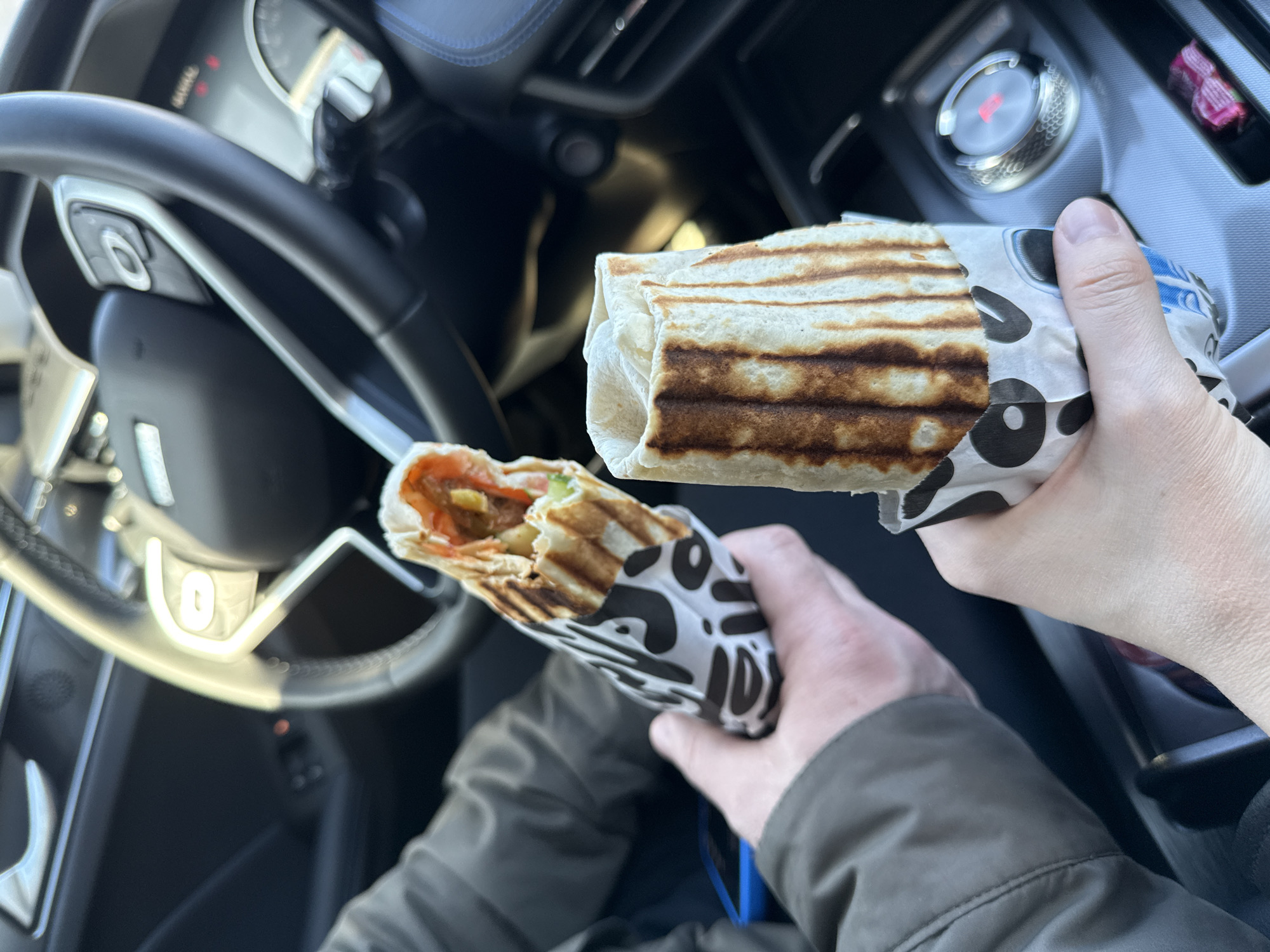 A person sits in the driver's seat of a car, holding two grilled wraps in paper packaging with a black and white pattern. One wrap is partially unwrapped, revealing fillings of tomato, lettuce, and other ingredients. The car's steering wheel and dashboard are visible. The car is most likely parked. The location information provided is incorrect, as there is no evidence to suggest the photograph was taken in Izhevsk, Russia.