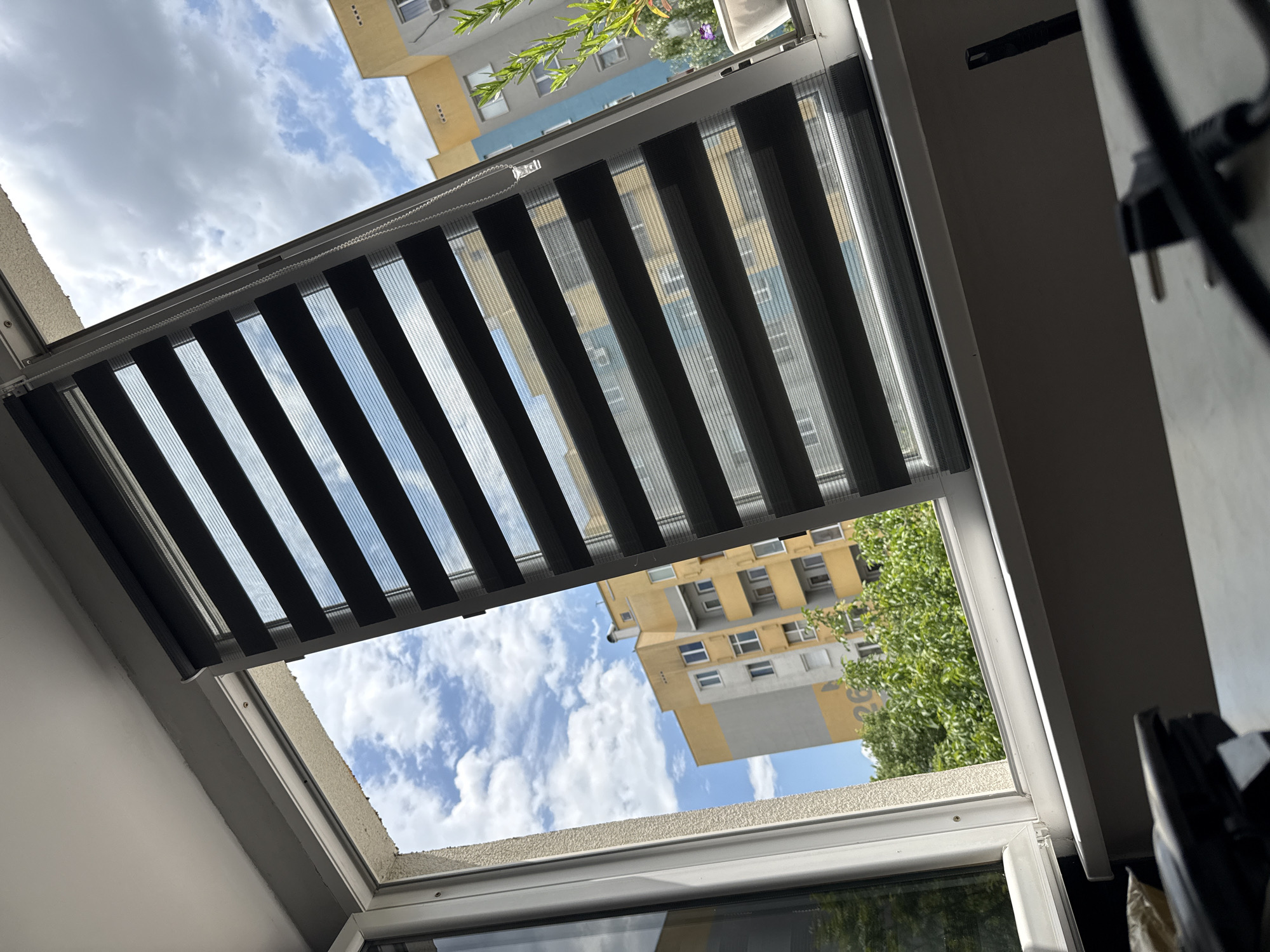 A low-angle, indoor shot shows a window looking out onto a view that includes buildings and a sky with clouds. The window has zebra blinds partially drawn, creating alternating stripes of light and shadow. Outside, parts of residential buildings are visible, with a yellow and white building on one side and a building with blue accents on the other. Some green trees are also present. The sky is mostly blue with fluffy white clouds. The window frame is white and is surrounded by a white wall on one side and a blurred interior on the other. The general location seems to be Stara Zagora, Bulgaria, based on the architectural style of the buildings.