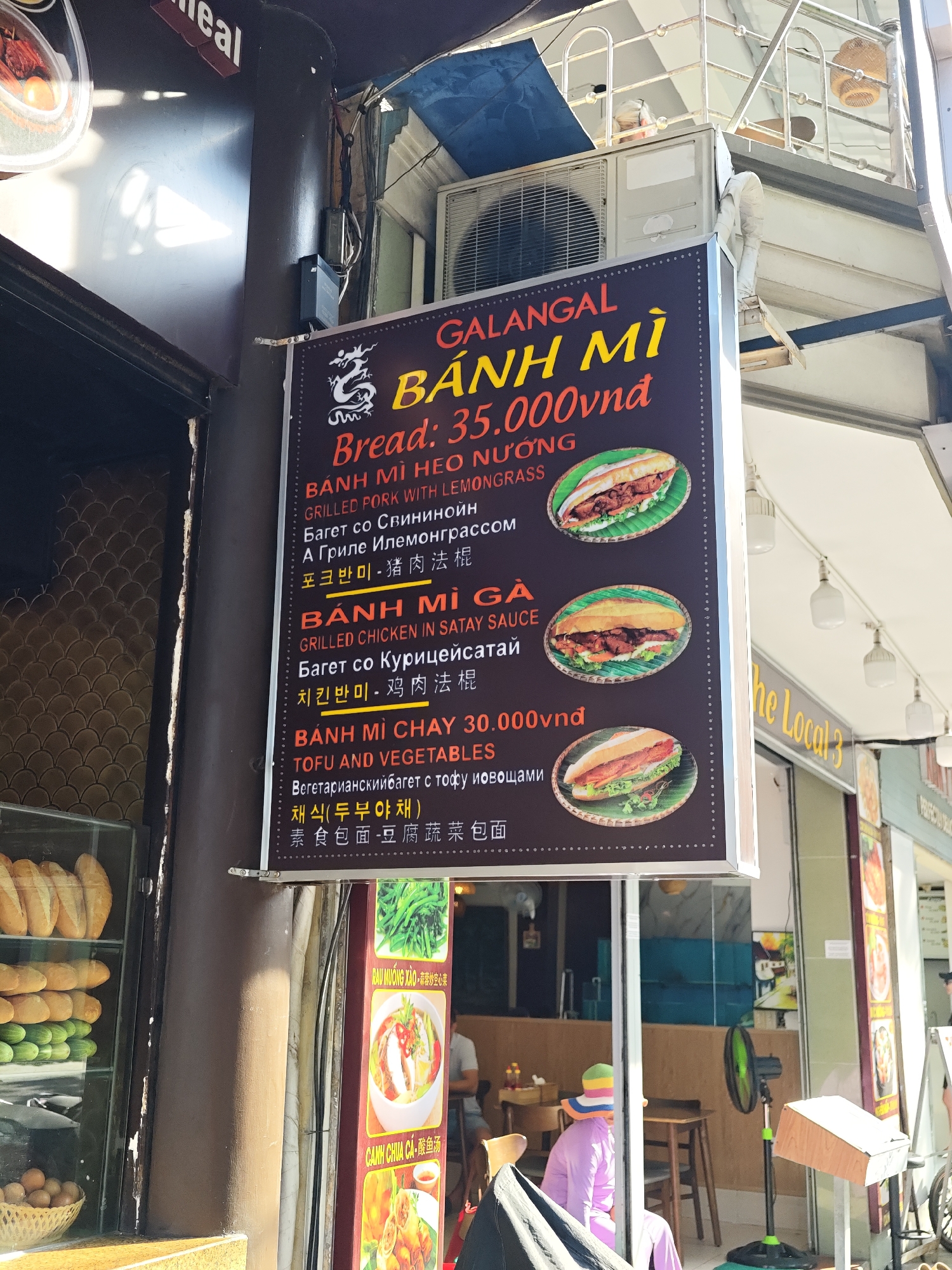 The image shows the exterior of a food establishment, likely a restaurant or food stall, in Nha Trang Ward, Vietnam. A prominent sign board, displaying menu items in Vietnamese, English, Russian, and Korean, is the central focus. The sign features images of banh mi sandwiches. The establishment appears to be situated on a street with other businesses visible in the background.
The sign advertises "GALANGAL BÁNH MÌ" with a price of "35.000vnd". It lists several banh mi options: "BÁNH MÌ HEO NƯỚNG GRILLED PORK WITH LEMONGRASS", "BÁNH MÌ GÀ GRILLED CHICKEN IN SATAY SAUCE", and "BÁNH MÌ CHAY 30.000vnd TOFU AND VEGETABLES". Below the English descriptions, there are translations in Russian and Korean, and Chinese characters are also visible.
To the left of the sign, a display window reveals a selection of bread loaves, cucumbers, and eggs, suggesting that these are ingredients for the food sold. The building itself has a slightly worn appearance, typical of many urban commercial areas. In the background, a sign that reads "the Local 3" can be seen.
The environment appears to be outdoors, with natural light illuminating the scene. The weather seems warm, as suggested by the light clothing of a person partially visible inside the establishment. A person wearing a purple outfit and a colorful hat is seated inside, facing away from the camera. A green fan is positioned near the entrance. The overall impression is of a bustling, everyday scene in a Vietnamese city.