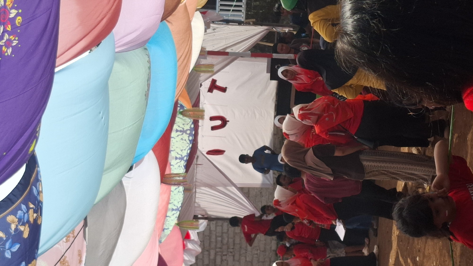 The image depicts an outdoor event under a canopy of colorful draped fabrics. In the background, a white tent structure with the red letters "OUT" visible on it is set up against a brick wall. Several people are gathered in front of and around the tent.
In the foreground and midground, a diverse group of individuals are present. Many are wearing red clothing, including shirts, hijabs, and headscarves. Some are interacting, while others appear to be observing the event. A young child in the lower left corner is wearing a red shirt with white and red face paint, and appears to be looking down and to the side.
The ground is dirt or packed earth. The overall atmosphere suggests a community gathering or celebration. While the exact location is not visually evident in the image itself, the prompt specifies it is in the city of Cilaku, Indonesia.