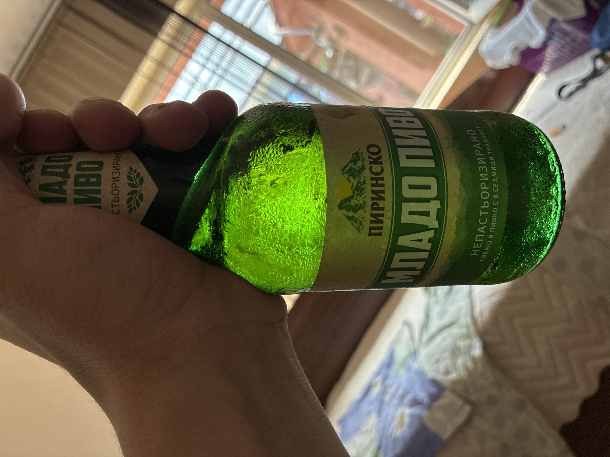 A close-up shot depicts a human hand holding a green glass beer bottle. The label reads "Младо Пиринско Пиво", which translates to "Young Pirinsko Beer", suggesting the beer is from the Pirin region. The bottle is covered in condensation. The background shows a blurred room interior with a window and a bed. The image does not provide explicit evidence to determine that it's located in the city of Varna, Bulgaria.