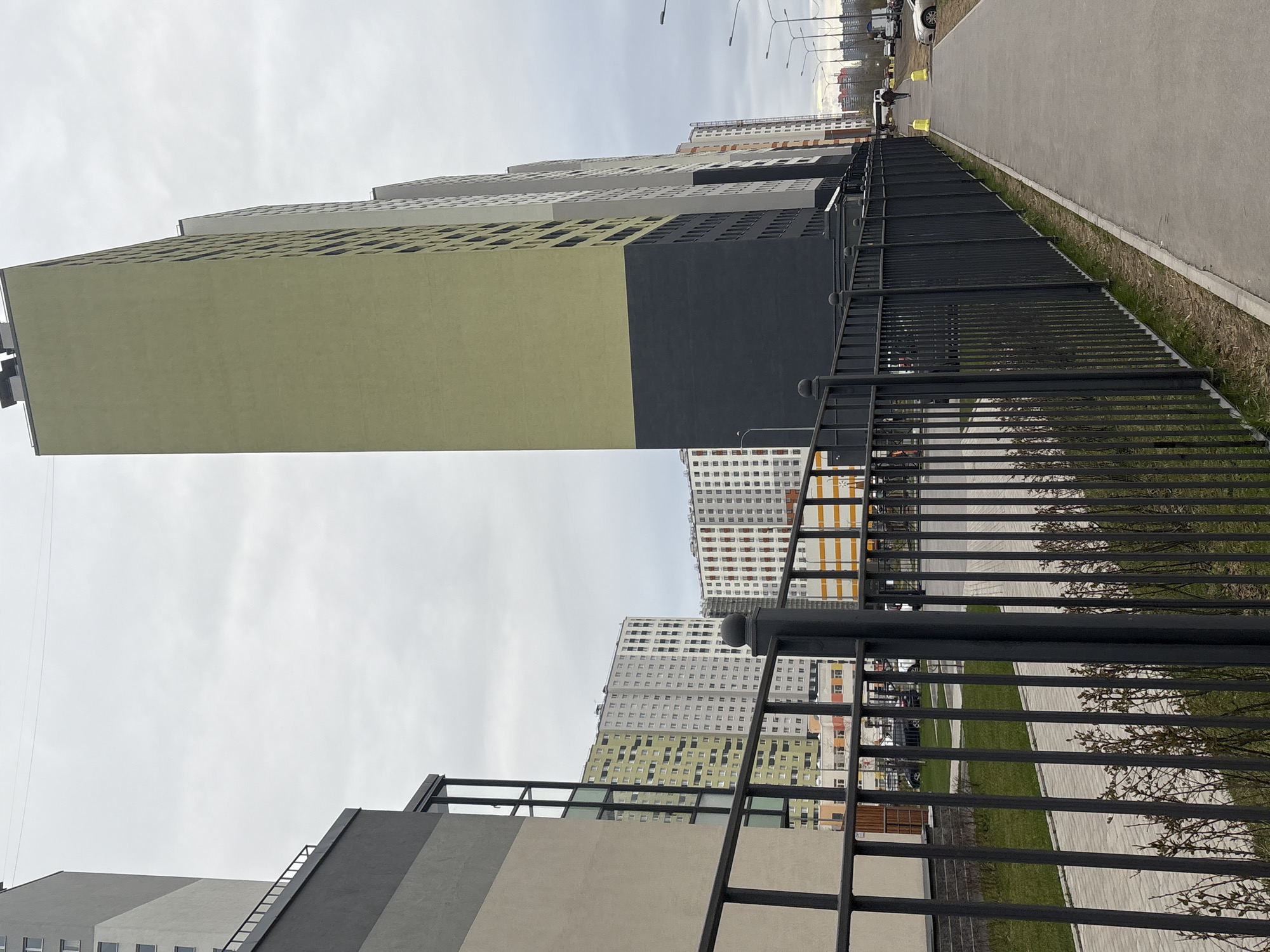 This image, rotated 90 degrees clockwise, captures a modern urban landscape in Saint Petersburg, Russia, under an overcast sky. The scene is dominated by tall, multi-story residential buildings, a black metal fence, and a paved pedestrian path.
In the foreground, a sturdy black metal fence with vertical bars runs diagonally across the frame, creating a strong visual line. Behind the fence, a paved path or sidewalk extends into the distance, flanked by thin strips of dry grass. Further back, a wider paved area, likely a road, hosts several light-colored vehicles, including cars and vans. A lone individual is visible walking along the paved path in the mid-ground.
The main subjects are the numerous contemporary apartment buildings that rise steeply into the sky. The most prominent building on the left features large, distinct sections painted in muted olive green and dark charcoal gray, with textured facades showcasing square window-like cutouts. Further along the street, a series of other residential blocks are visible, displaying varied color schemes including white, light gray, and splashes of orange and lighter green, indicating a large-scale housing development.
The time of day appears to be daytime, but the uniformly overcast sky suggests cloudy weather, lending a muted light to the scene. The overall impression is one of a functional and newly developed residential area. No discernible text is visible within the image.