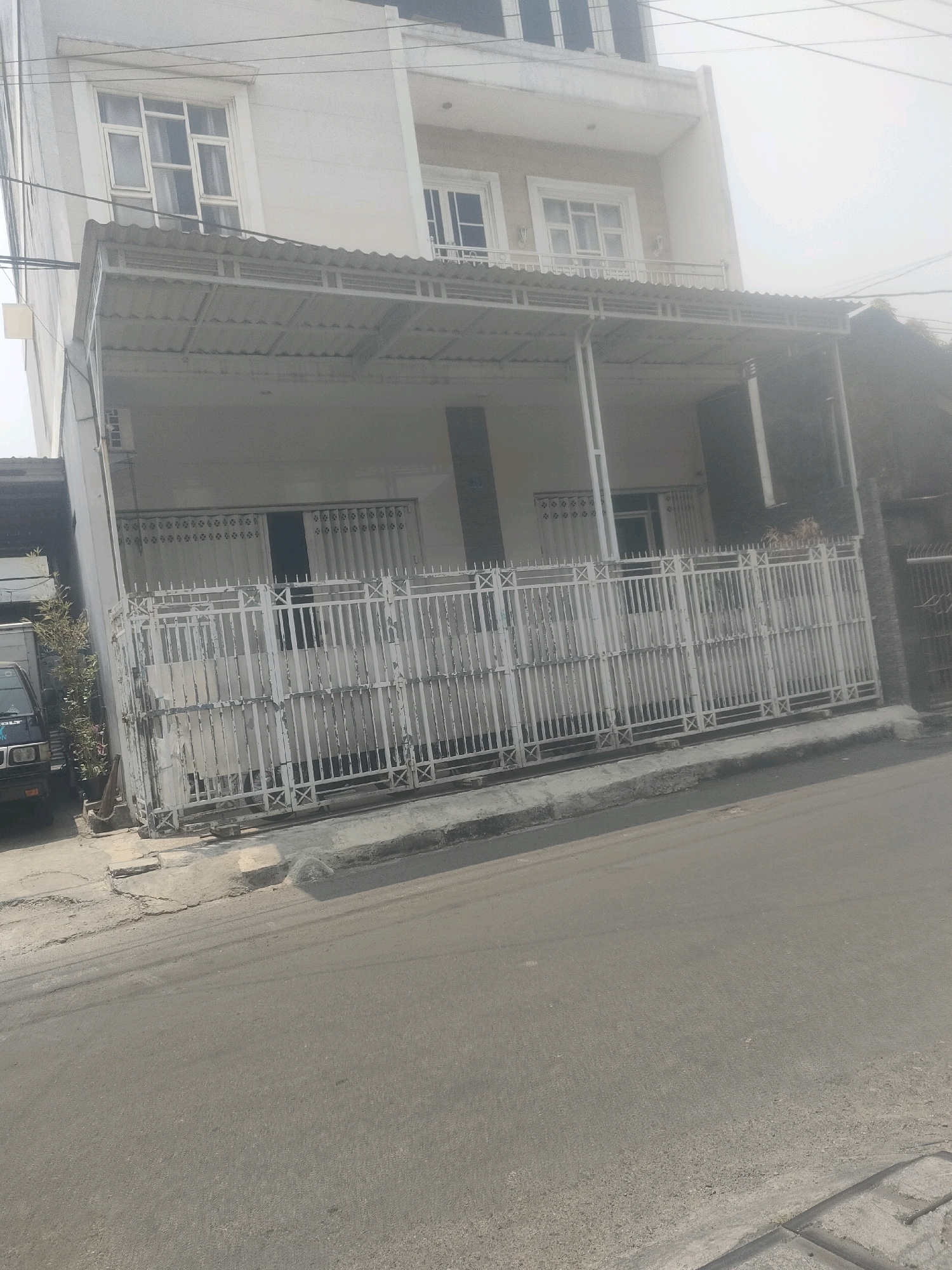 This is a photograph of a residential building with a white facade and a corrugated metal awning extending from the second story. The building is located on a street in Surabaya, Indonesia. A white metal fence encloses the property, and a dark-colored vehicle is parked to the left of the image, partially obscured by the fence and some greenery. The street in front of the building is paved and appears to be in moderate condition, with some cracks and unevenness visible. The sky is overcast and bright, suggesting a daytime scene.