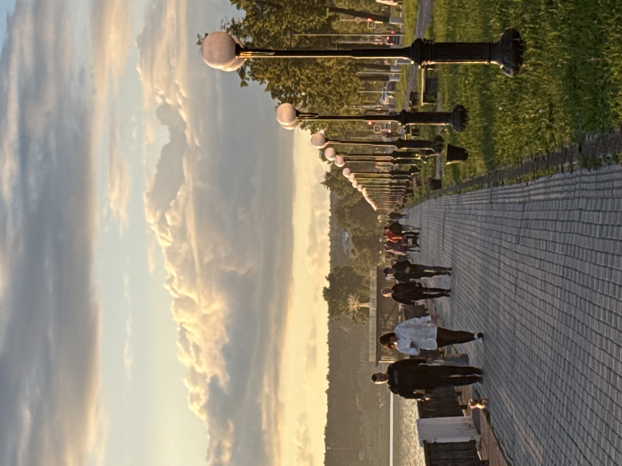 This media file captures an outdoor scene at either sunrise or sunset, characterized by soft, warm light. A wide, paved pathway, constructed from small rectangular paving stones, extends diagonally from the foreground towards the background on the right side of the image.
Along the left edge of this pathway, a long, receding line of ornate black lampposts with globe-shaped white lights creates a strong sense of perspective. Beyond the lampposts, there's a strip of green grass followed by a dense line of trees, which appear to border a body of water or a lower landscape that is partially visible on the far left.
Several people are walking along the paved path, engaged in leisurely activity. In the midground, one individual in a white shirt and dark pants walks away from the viewer, while another in a dark jacket is closer to the foreground. Further down the path, multiple figures, some solitary and others in small groups, can be seen strolling. Their attire is casual, suggesting a recreational outing.
The sky dominates the upper left portion of the image, showcasing a dramatic cloud formation. Some clouds are brightly illuminated with golden and orange hues, indicating the low angle of the sun, contrasting with areas of darker, greyish cloud cover. This warm, directional lighting strongly suggests either early morning or late afternoon. The overall atmosphere is peaceful and serene. There is no visible text in the image. The setting appears to be a public promenade or embankment, consistent with a scenic area in Votkinsk, Russia.