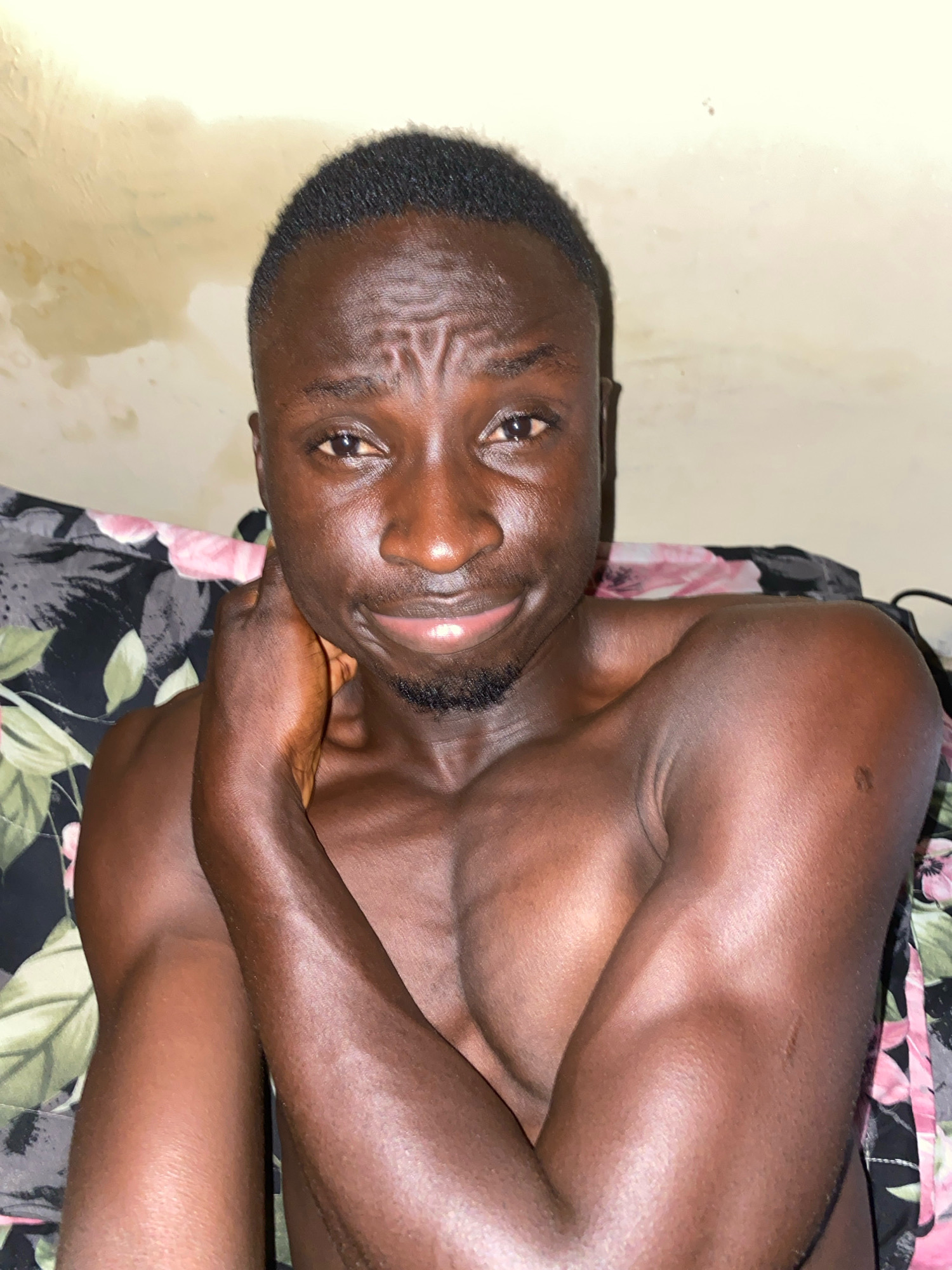 The media file is a close-up, selfie-style photograph of a shirtless Black man. He is lying down, with his head resting on a pillow with a floral pattern featuring pink and green flowers on a dark background. The man's face is the primary focus, with his left arm bent and his hand resting on his neck. He has short, dark hair and a small beard. His expression is a slight smile, and his eyes are looking directly at the camera. The background wall is a light, creamy color and appears textured. There are some smudges or water stains on the wall behind him. The lighting suggests an indoor setting, possibly at night or in a dimly lit room, given the warm tones and shadows. The overall mood is relaxed and intimate. No visible text is present in the image.