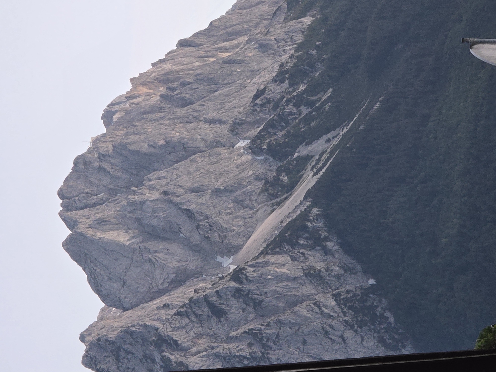 The image shows a majestic mountain peak in Garmisch-Partenkirchen, Germany, bathed in soft, diffused light. The mountain's rocky slopes are rugged and textured, with visible striations and crevices. Patches of snow are scattered across its face, contrasting with the gray rock. The lower slopes are covered in a dense, dark green forest that transitions into the rocky terrain above. A small, white building or structure is visible near the summit, perched on a rocky outcrop. The sky above is a pale, clear blue. The foreground features a blurred dark object on the left, likely a railing or support, and a portion of a white satellite dish or similar object at the bottom right, suggesting a vantage point from a building or observation deck.