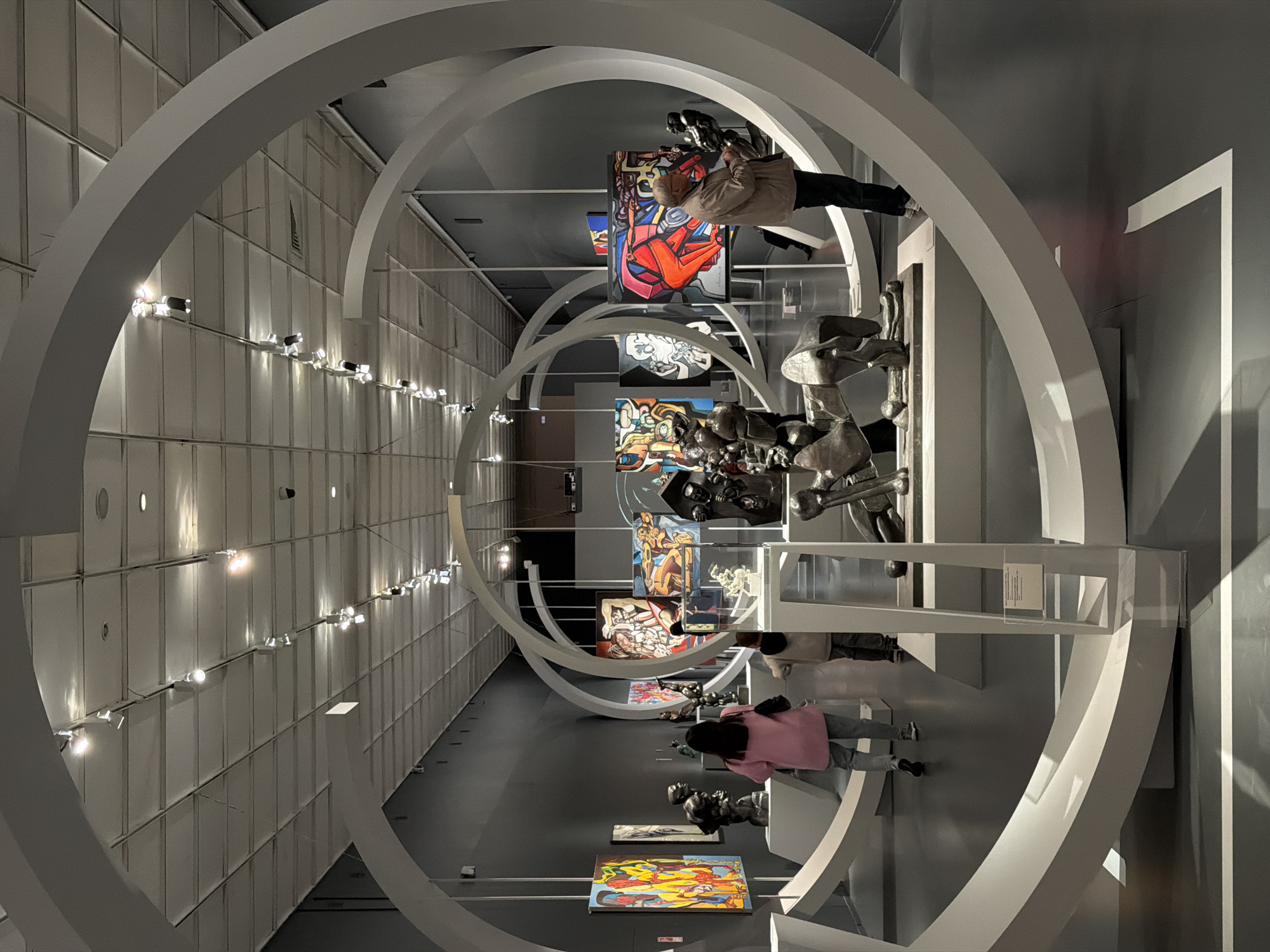 The image shows an interior view of an art gallery. The space is characterized by a series of white arches that form a tunnel-like structure. Paintings and sculptures are displayed throughout the gallery. Several people are present, including a person in a tan jacket taking a photograph of a painting, and another person wearing a pink shirt. The walls are a combination of white and grey, and lighting fixtures are visible along the walls. The gallery has a modern and spacious feel. The scene takes place in the Zurab Tsereteli Art Gallery, located in Moscow, Russia.