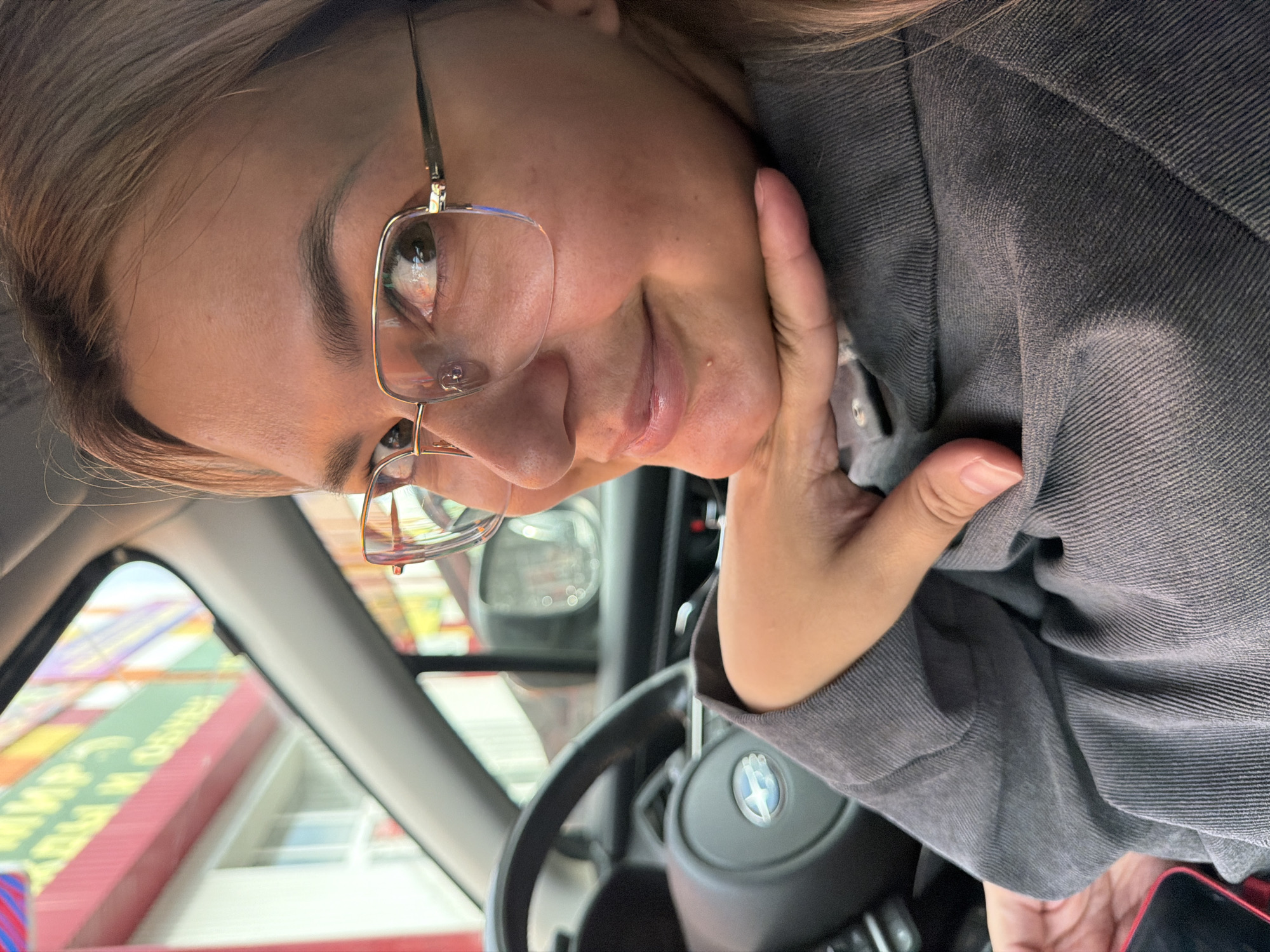 A medium close-up shot shows a woman with blonde hair, wearing glasses and a gray shirt, leaning her chin on her hand while sitting in the driver's seat of a car. The steering wheel is visible on the left side of the frame. The background, seen through the car window, displays a colorful building with red and green accents, along with signage that suggests an urban environment. The lighting is bright, indicating it is daytime. The photo was taken in Birobidzhan, Russia.