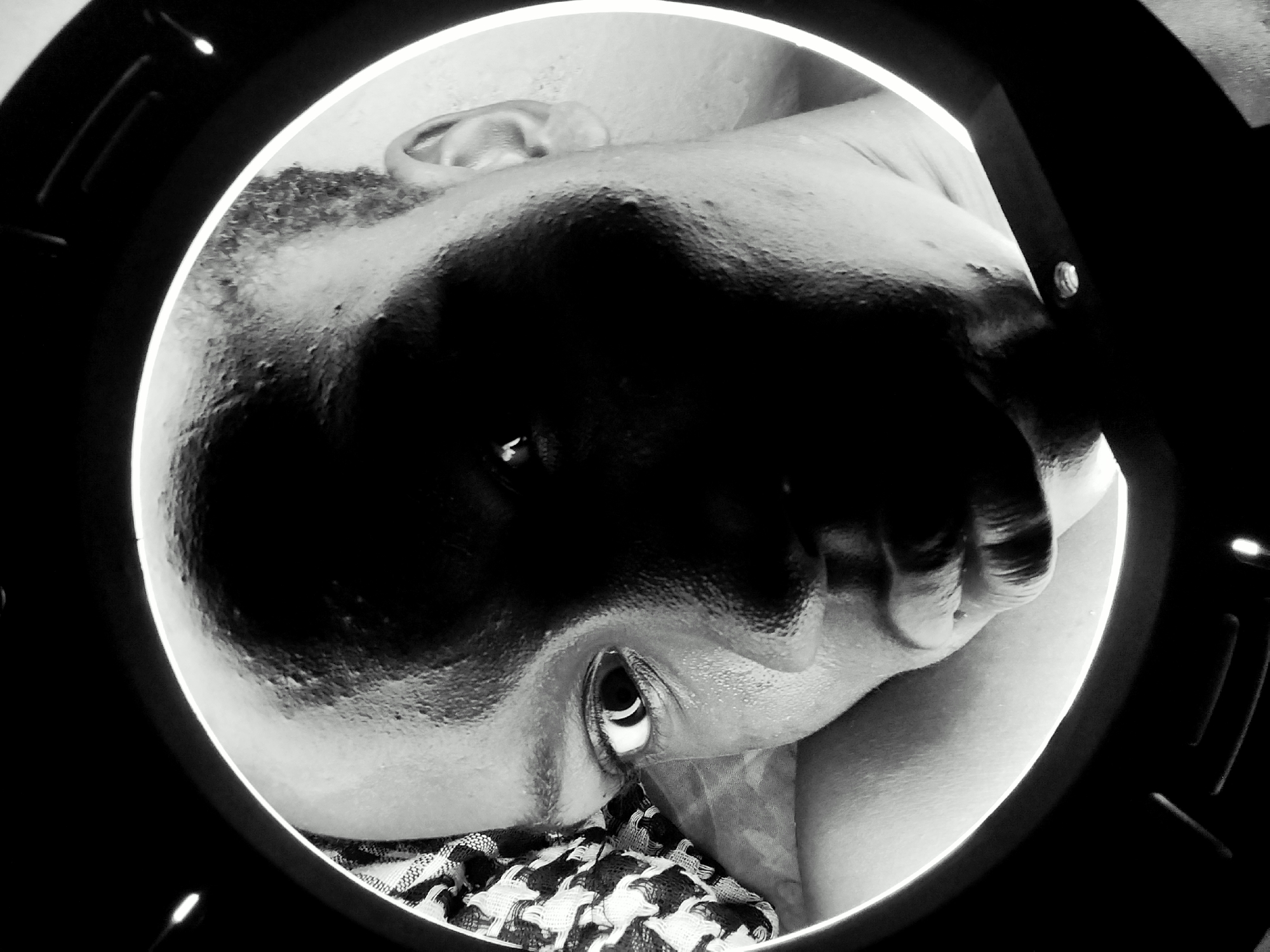A close-up, black and white image presents a person's face framed by a circular object. The person appears to be looking upwards, with one eye clearly visible. Their hand is positioned near their face. A patterned fabric is visible beneath the person's face. The circular frame has a thicker outer ring. The location is Unknown, Unknown.
