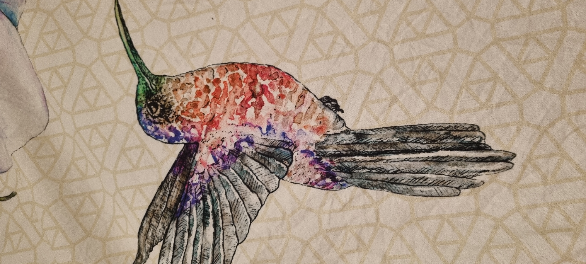The image is a close-up of a fabric print featuring a hummingbird. The hummingbird is depicted in a watercolor-style illustration, with its body displaying a vibrant mix of colors, including red, orange, purple, and green. Its wings are spread, showing detailed feathering in shades of gray and blue, with purple accents. The bird has a long, thin beak, colored green. To the left of the hummingbird, a portion of a light purple, floral-like shape is visible, suggesting it might be part of a larger floral design. The background of the fabric is a light, neutral color with a geometric pattern of intersecting lines forming a grid of triangles and quadrilaterals in a slightly darker, beige tone. The image captures the artistic rendering of the hummingbird and the textured pattern of the fabric. No text is visible in the image.