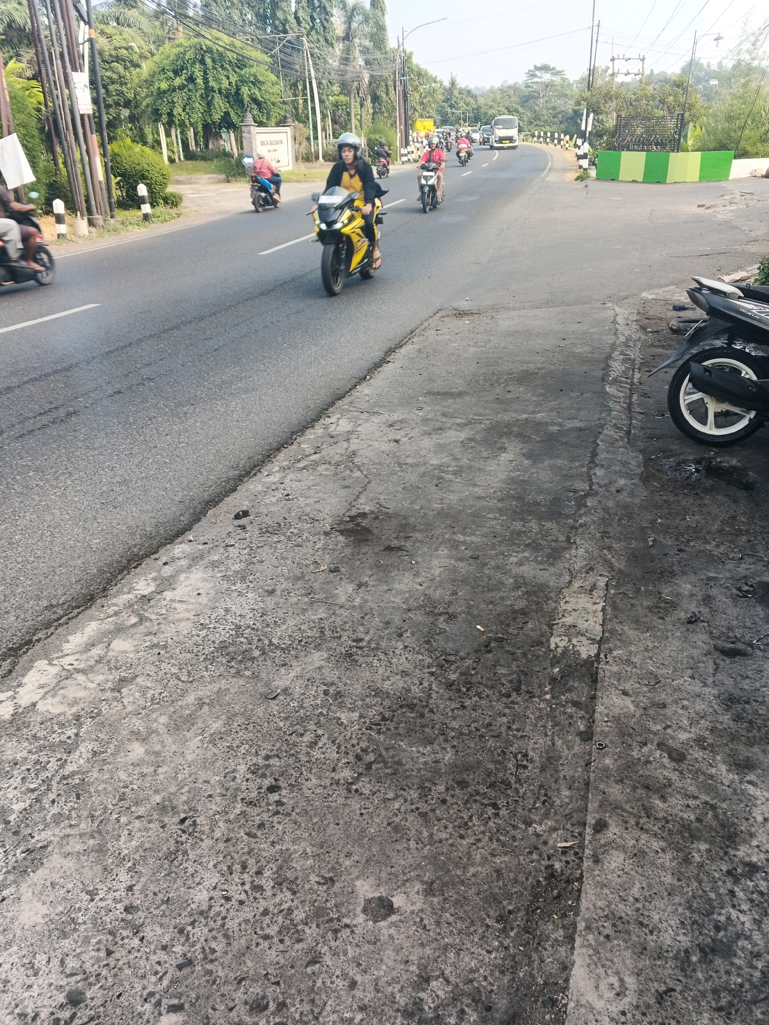 The image depicts a street scene in Ungaran, Indonesia, likely on a sunny day. In the foreground, the rough, grey asphalt of a sidewalk is visible, showing signs of wear and tear, with dark stains and debris scattered across its surface. A portion of a parked black motorcycle with a white rim is visible on the right side of the frame. The road curves to the left, with several people riding motorcycles.
On the road, a person in a yellow jacket and black helmet is riding a yellow and black sports motorcycle, heading away from the viewer. To their left, another person on a motorcycle is also traveling in the same direction. Further down the road, more motorcycles are visible, suggesting traffic.
The background features a lush green environment with trees lining the road. A white truck is seen in the distance, navigating the curve. There are utility poles and wires present, typical of urban infrastructure. A sign with text "MELTA BALEMUR" is visible on a building to the left, and a white and green barrier is present near the road's edge. The overall impression is a busy, active street in a semi-urban area of Ungaran.