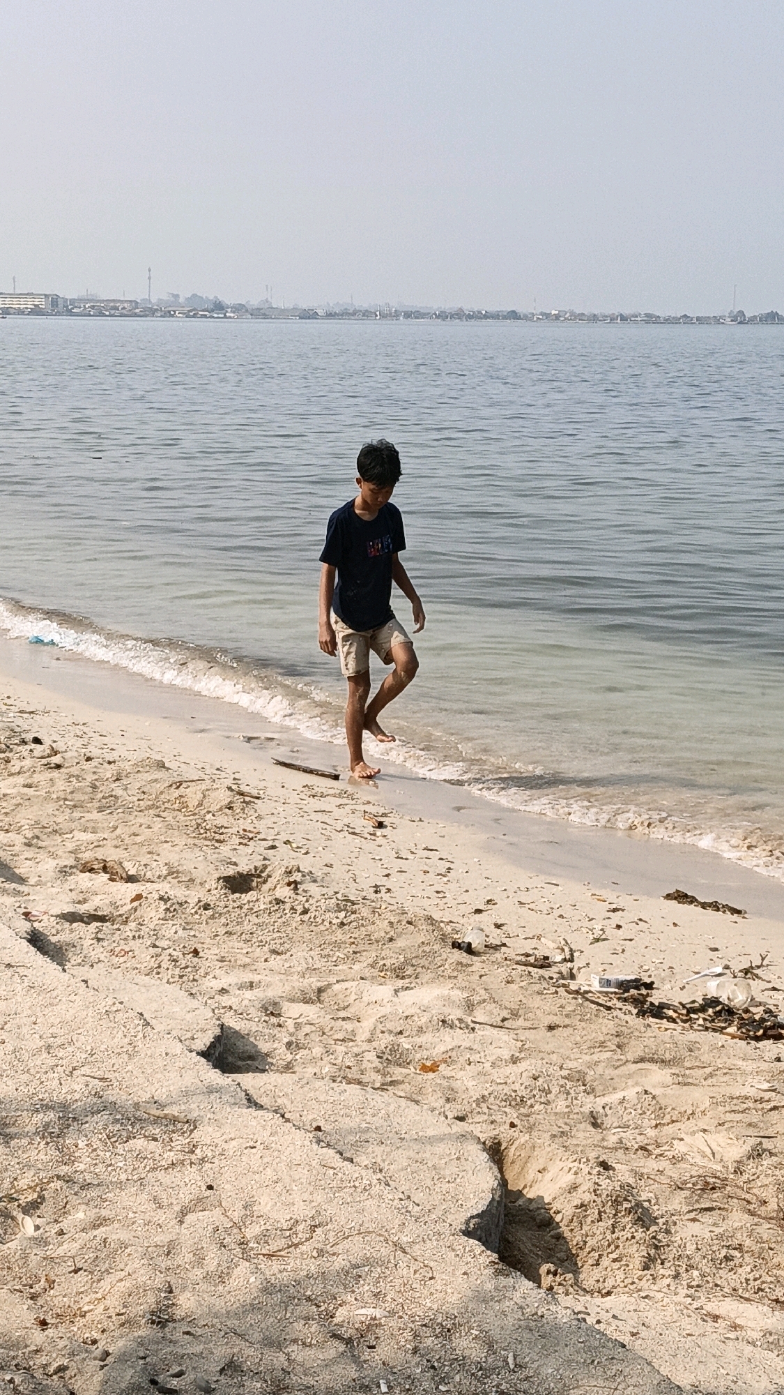 A young boy walks along the edge of the water on a sandy beach. He is wearing a dark t-shirt and light-colored shorts. The water is calm and appears shallow, with small waves lapping at the shore. The beach is covered in sand and debris, including a few pieces of trash. In the background, a hazy cityscape is visible across the water. The location is Cikal, Indonesia.